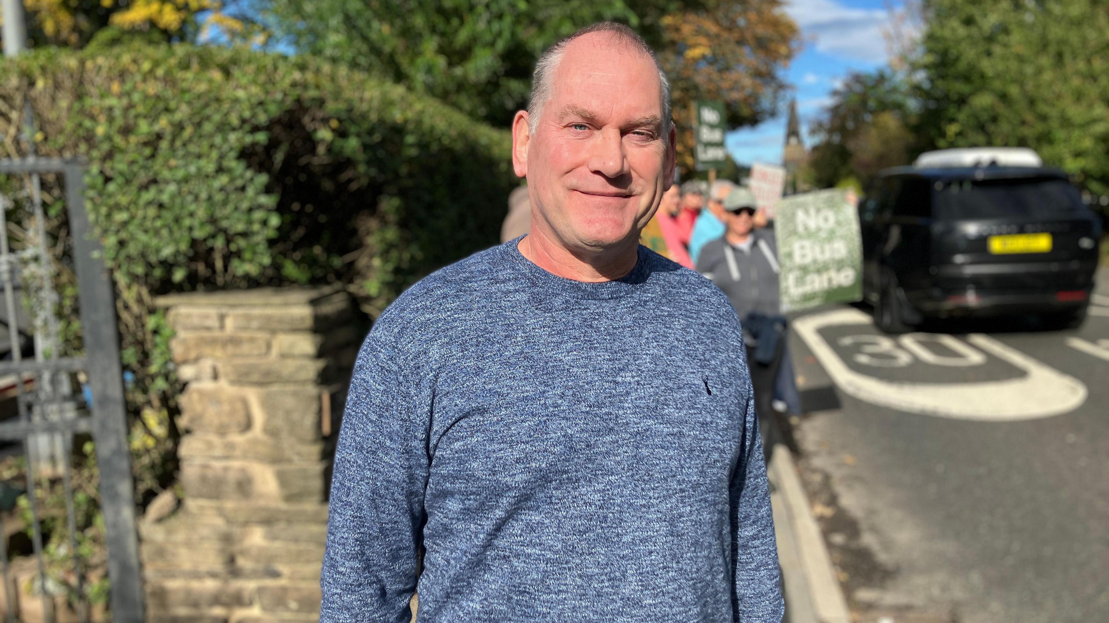 A man in a blue jumper with white hair standing at the side of the road with protesters. There is a hedge behind him and a black Range Rover-type car to the side. People hold placards behind him.