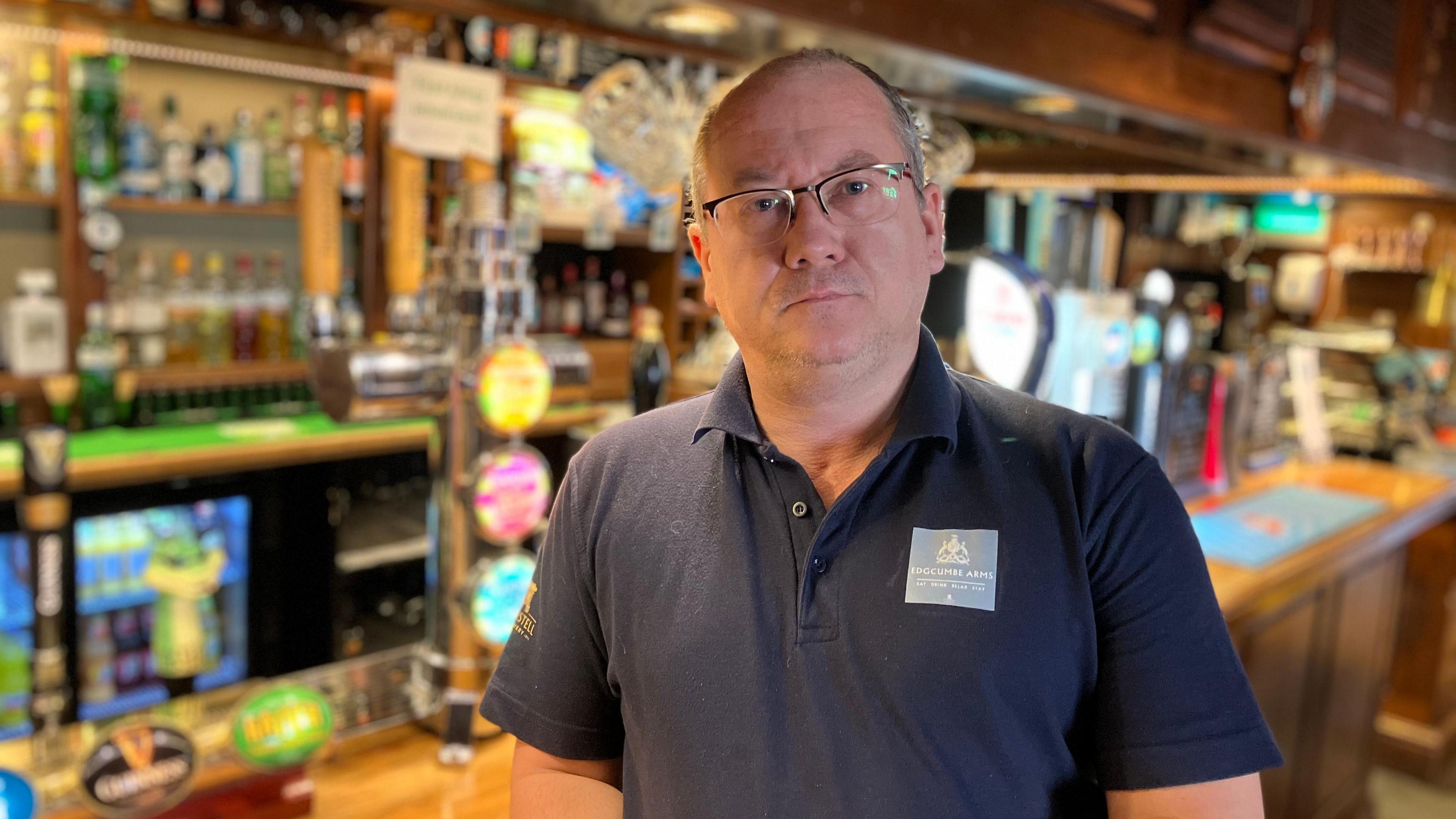 A man in a black t-shirt stands in front of a brightly lit bar in a pub. 