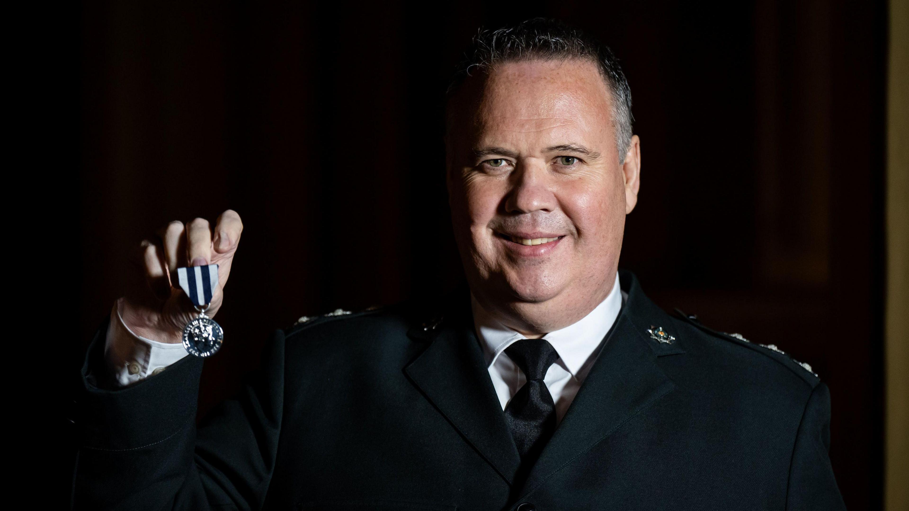 John Caldwell holds up his King's Police Medal while smiling at the camera.