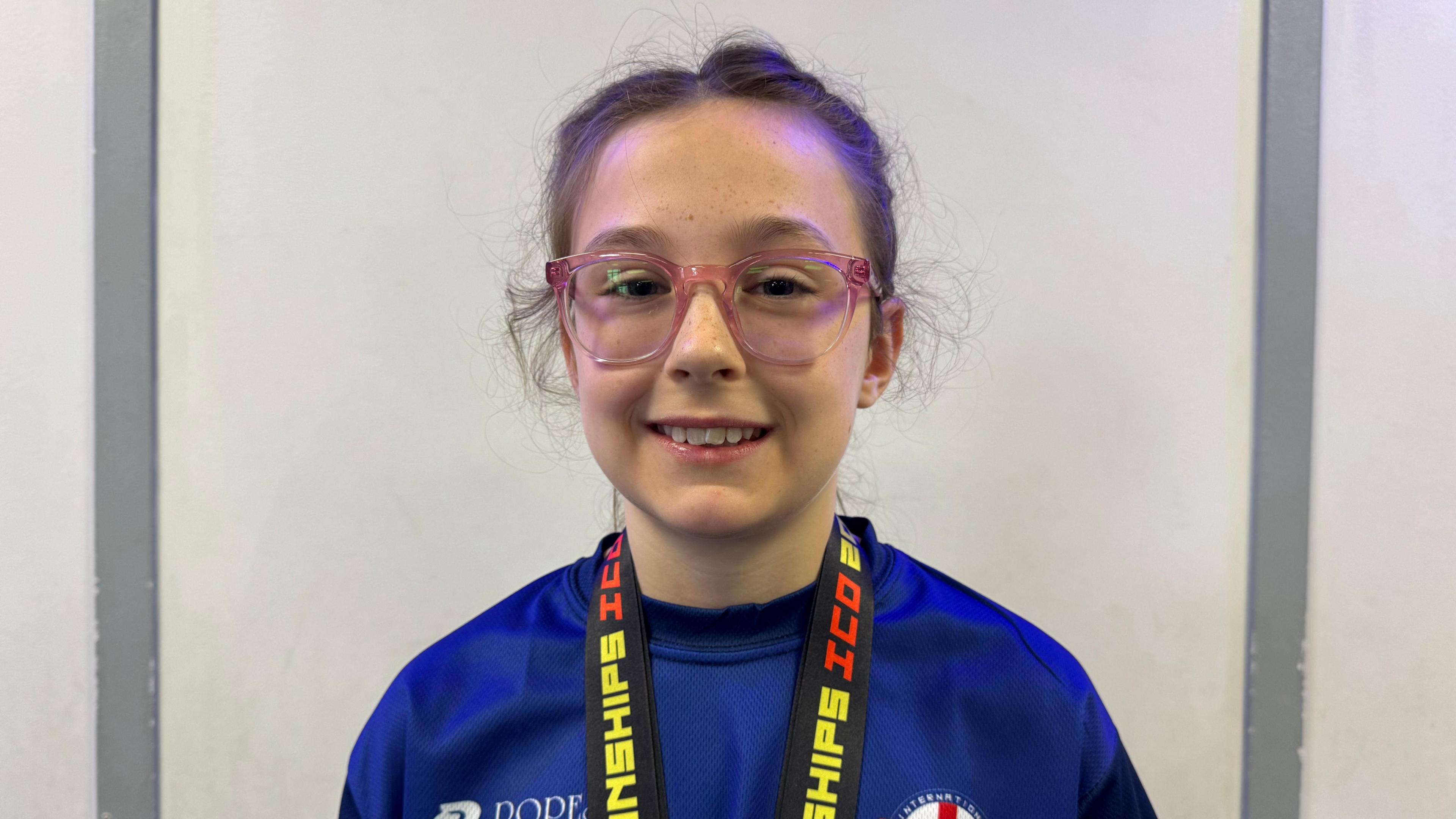 A young girl wearing a blue T-shirt and pink glasses has two medals around her neck