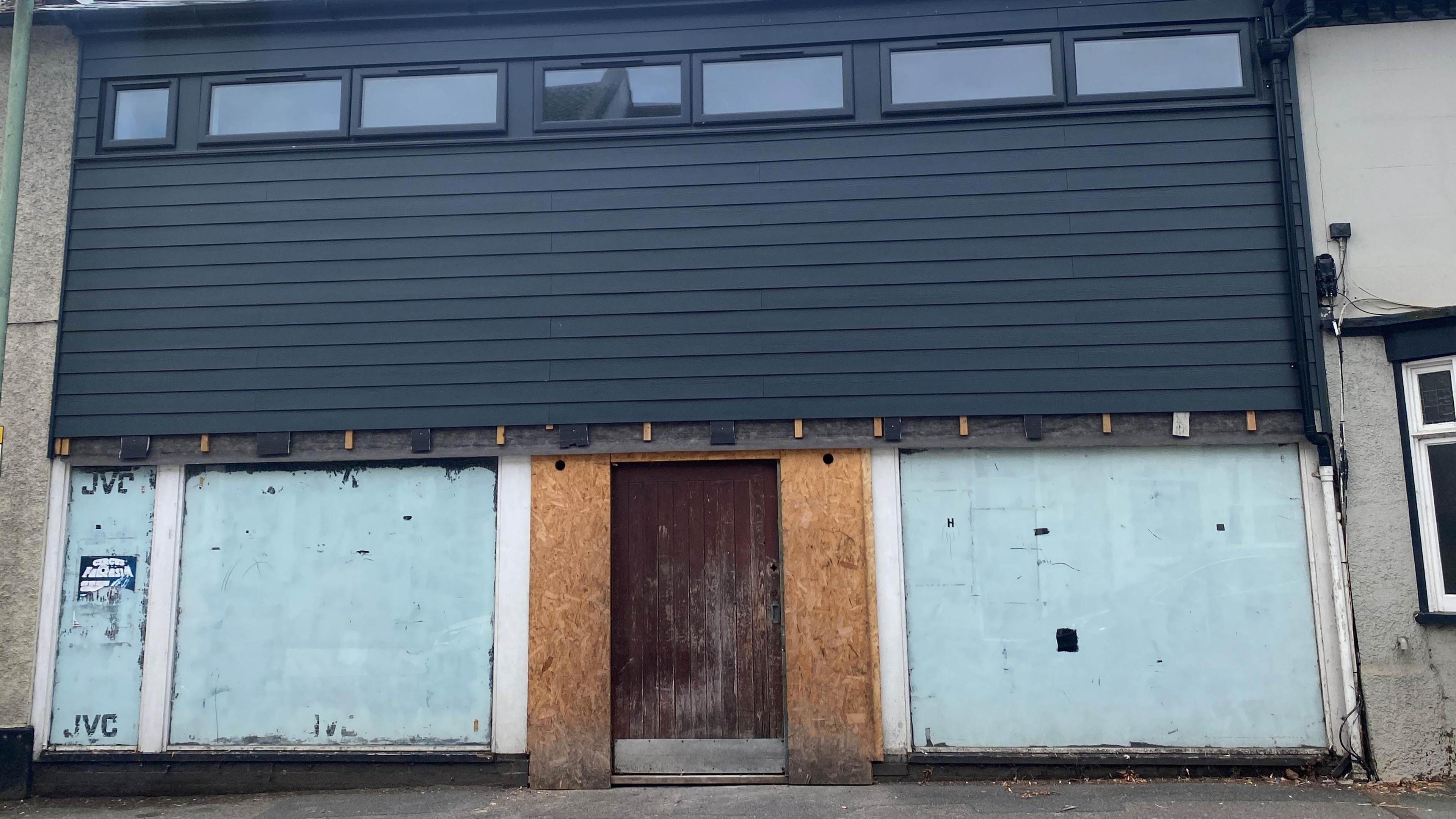 A two-storey building with black panels at the top of the building and the bottom two windows and doors are boarded up.