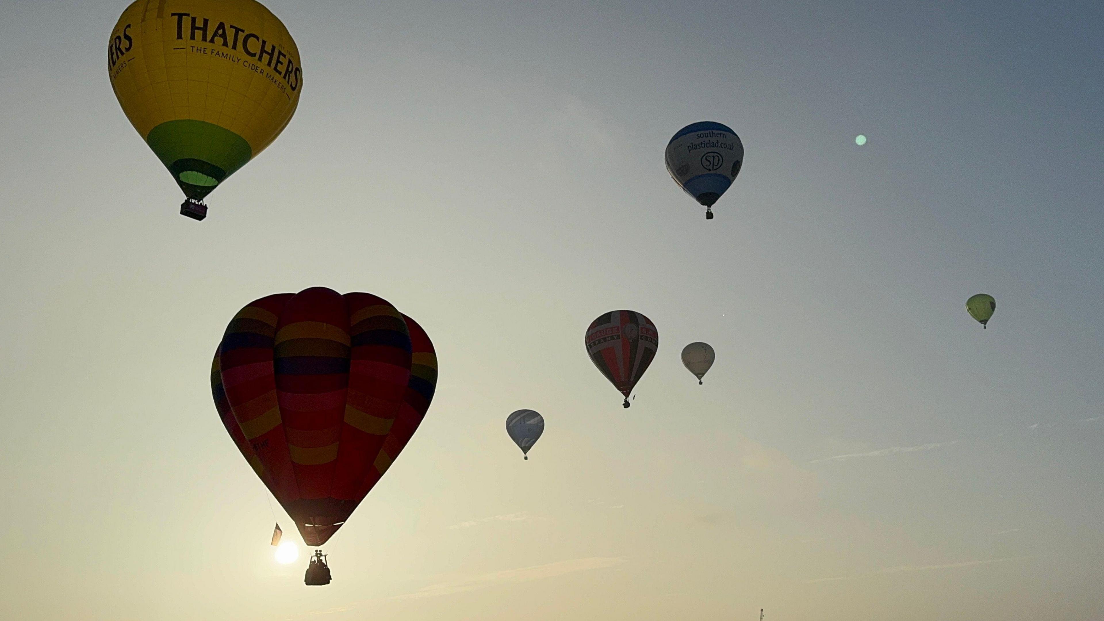 Several hot air balloons in the sky. They are all different sizes in the photo due to them being at different heights and distances. Most of them are silhouetted by the morning sun.