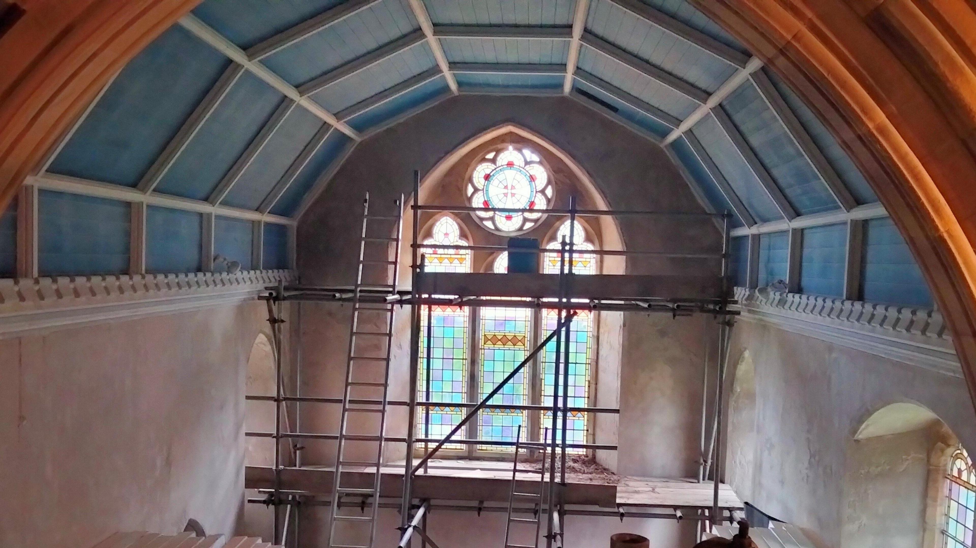 The inside of a church with blue ceilings, white washed walls and stained glass windows. Inside scaffolding and ladders shows construction work underway.