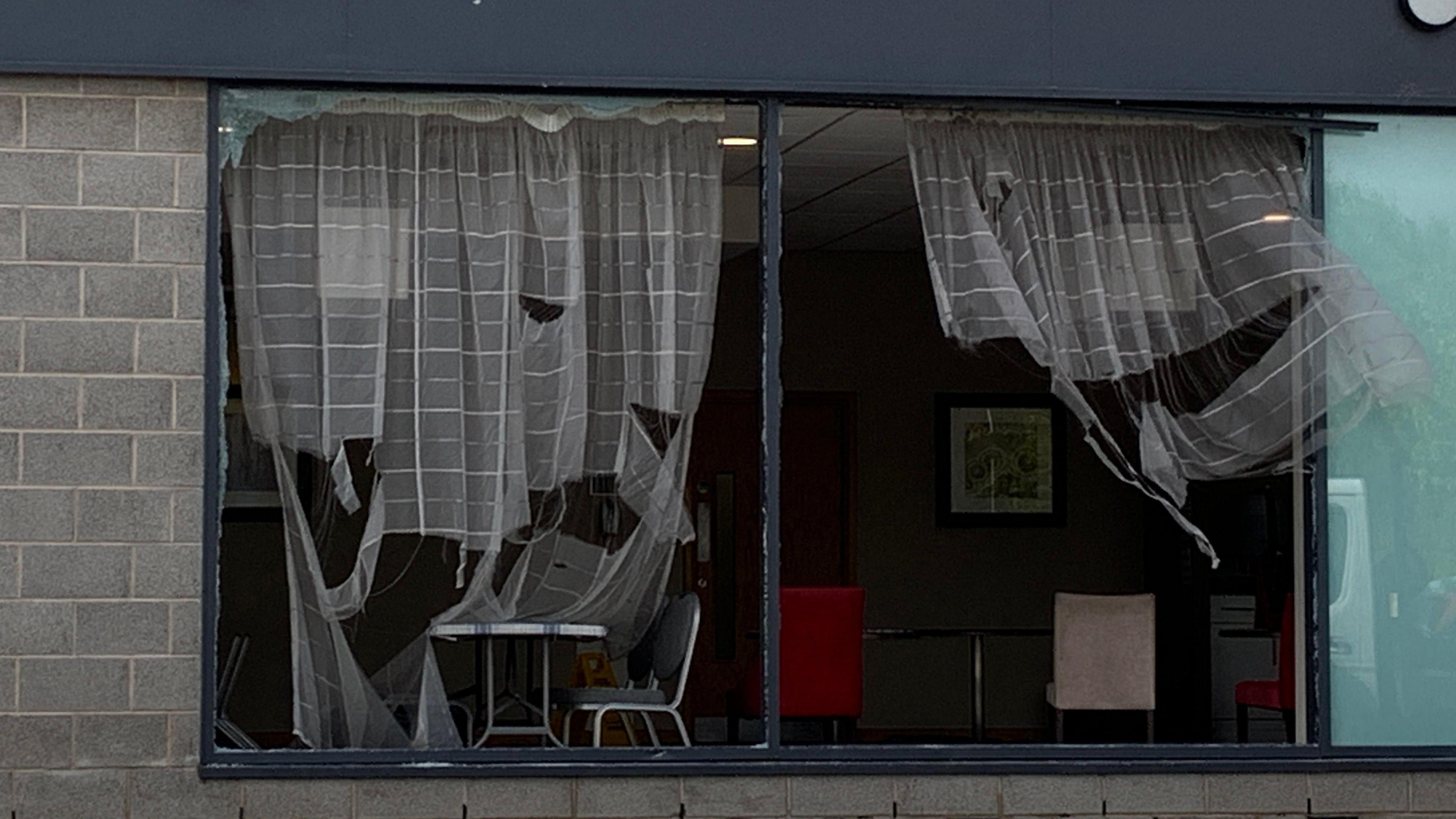 A broken window shows furniture such as tables and chairs behind ripped white curtains.