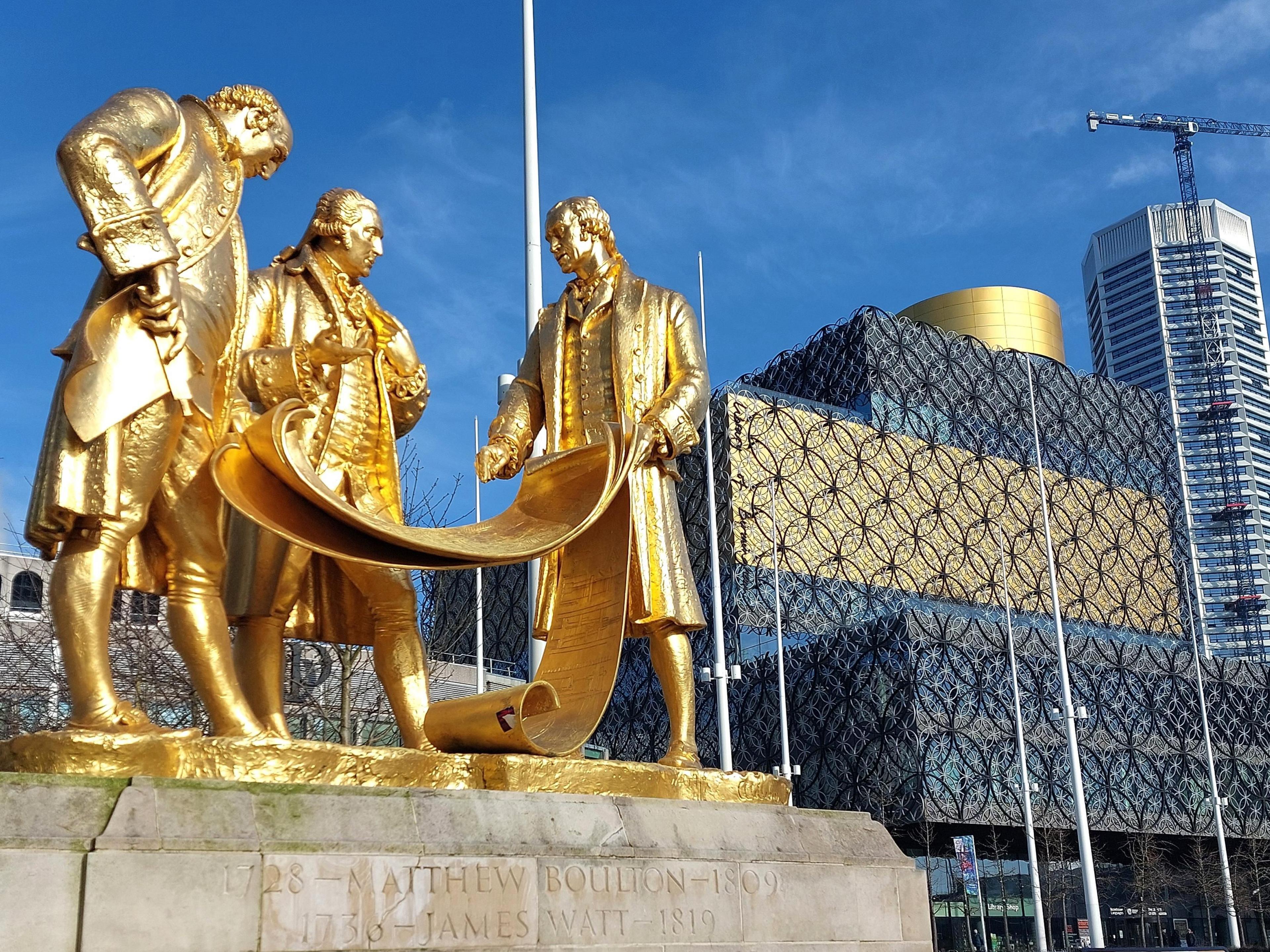 A vibrant golden statue of three men apparently discussing the contents of scrolls unrolled in front of them. It stands on a plinth of Portland stone, with Birmingham's modern central library behind, under a blue sky.