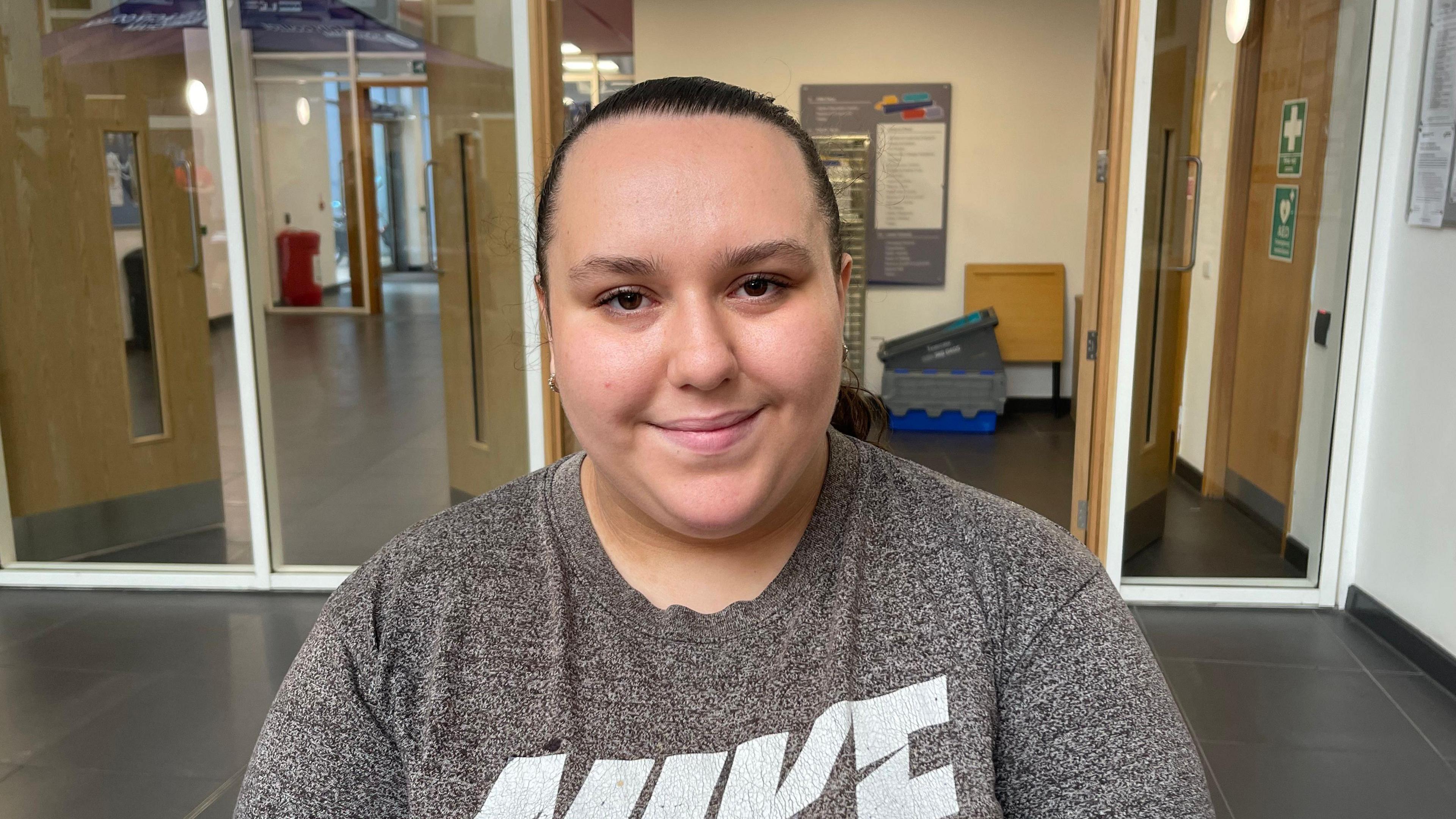 Bethany Clarke, a young woman, is standing in a school hallway. She's wearing a grey Nike T-shirt and has her hair tied back in a ponytail. She is smiling at the camera. 