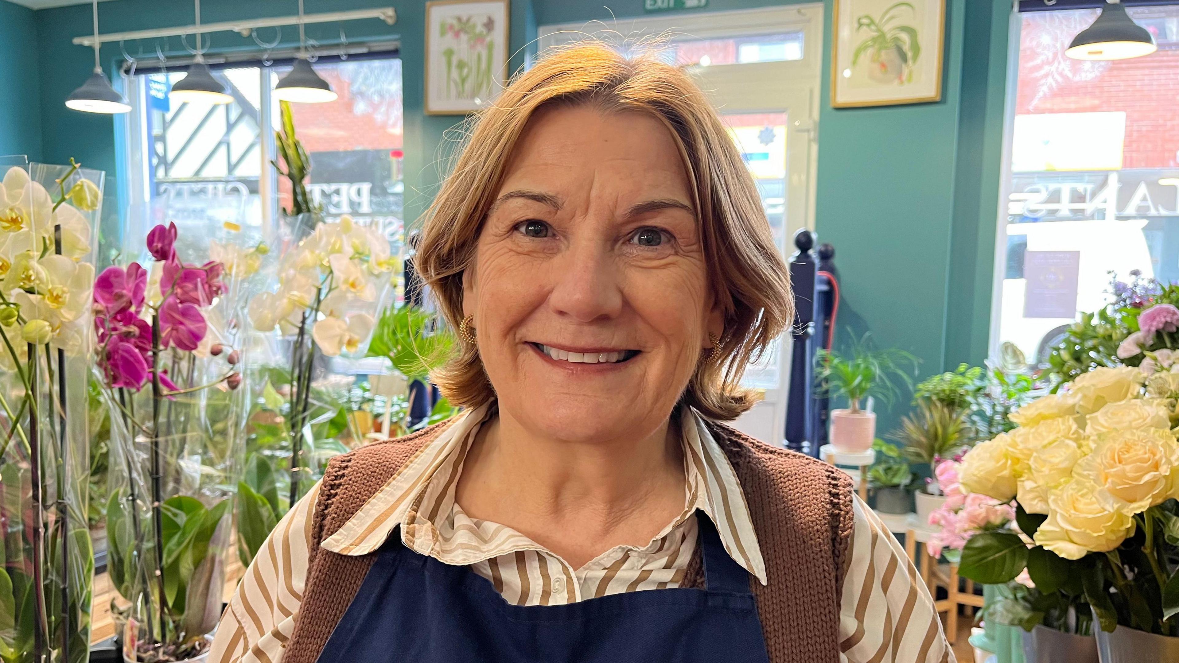 Jean pictured looking into the camera and smiling. She has short light brown hair and is wearing a brown and white striped shirt with a brown sleeveless cardigan and a navy apron. On tables around her are orchids, roses and houseplants. The walls of the room are teal blue and there are four windows on the far wall.