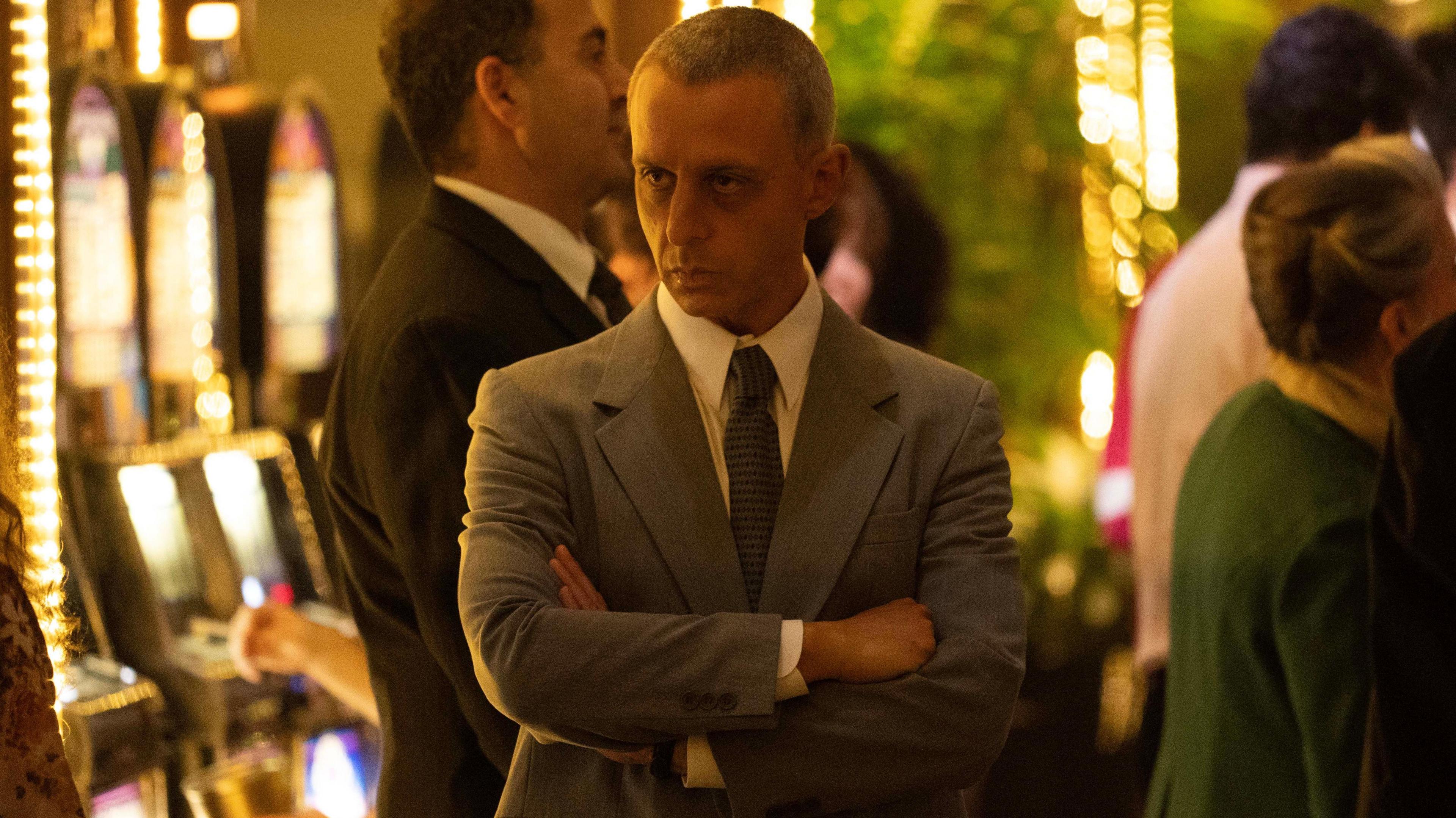 Jeremy Strong as Roy Cohn in The Apprentice - he is surrounded by slot machines and is wearing a grey suit and has his arms crossed