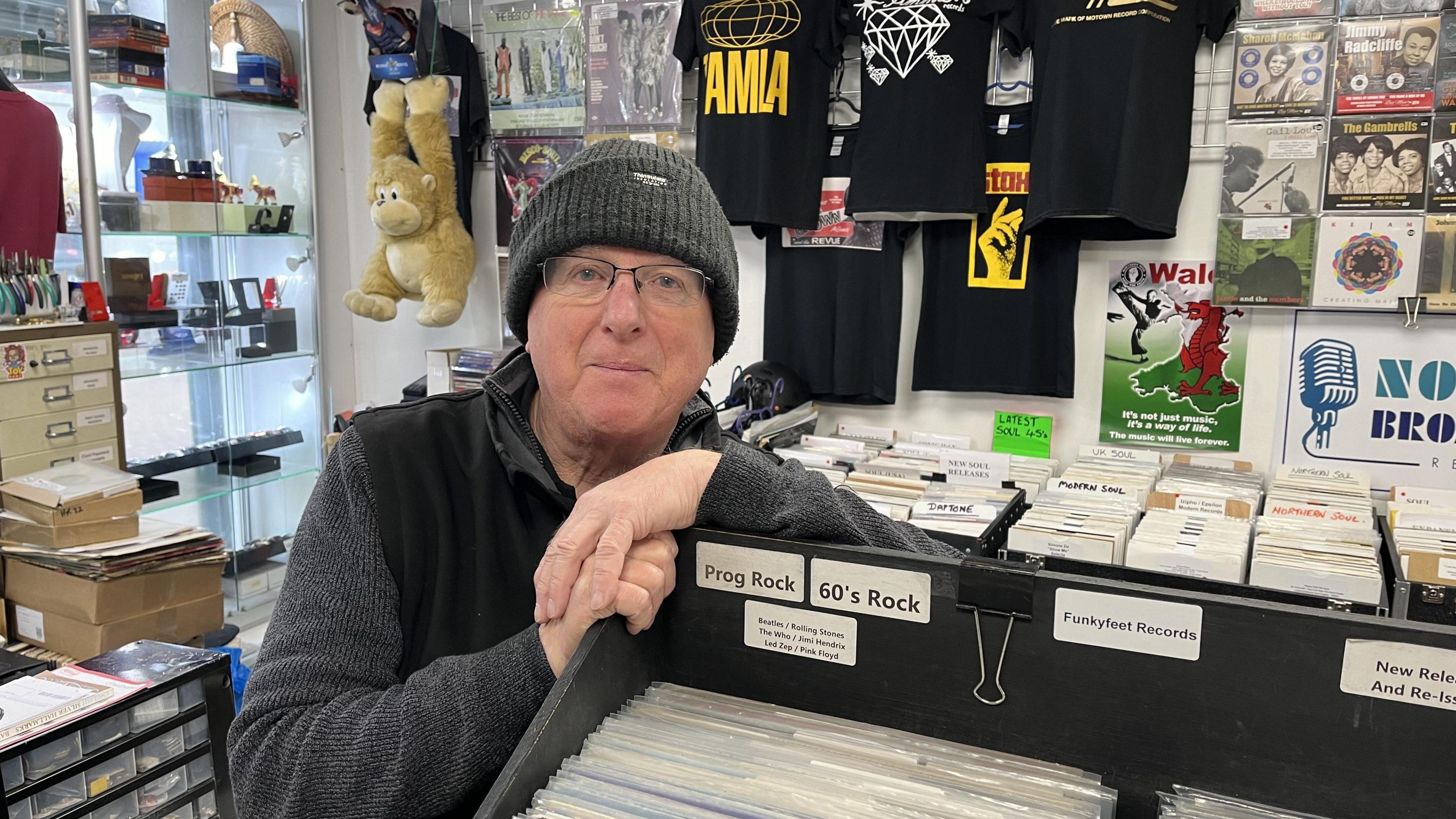 Mark Speakman wearing a grey hat in his record shop and leaning on one of the many racks of 12 inch vinyls. 