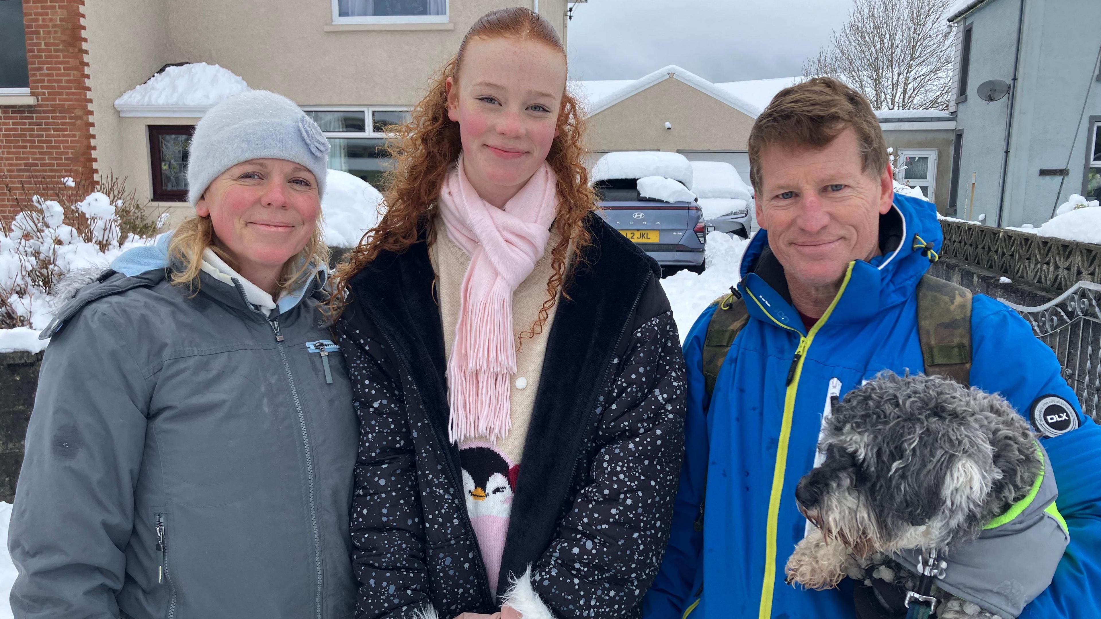 Helen Mcloughlin, Ela Mcloughlin, 14, Conan Blake and dog Madog, wearing winter gear, pose and smile outside a snow-covered home and car.