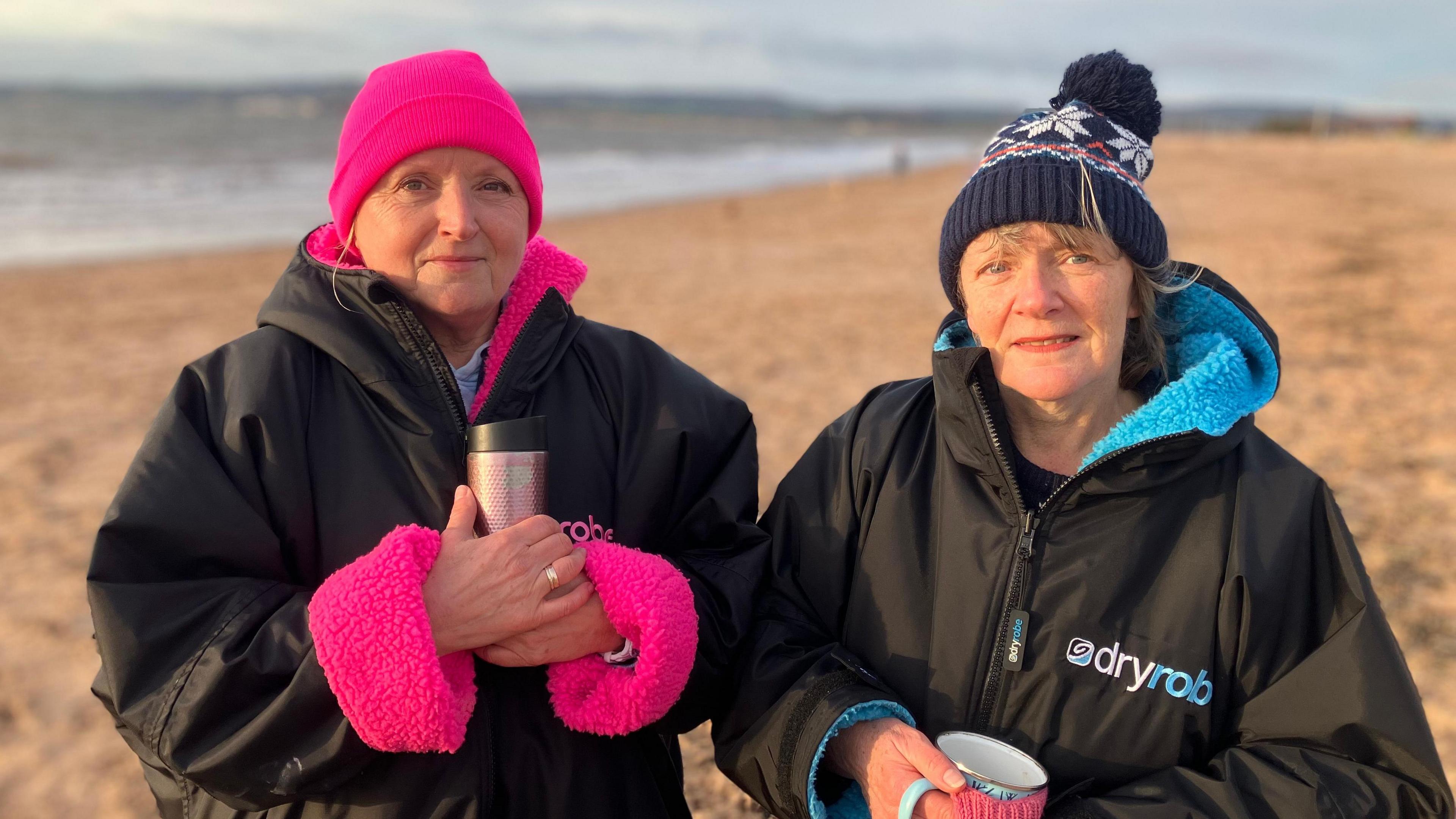 Janet Penrose (left) and Marcia Fletcher (right) stand on Exmouth beach wearing pink and blue fleece-lined Dryrobe jackets respectively while holding onto mugs. both are wearing wearing woolly hats - Janet has a pink one and Marcia a dark blue hat with a white star pattern on it.