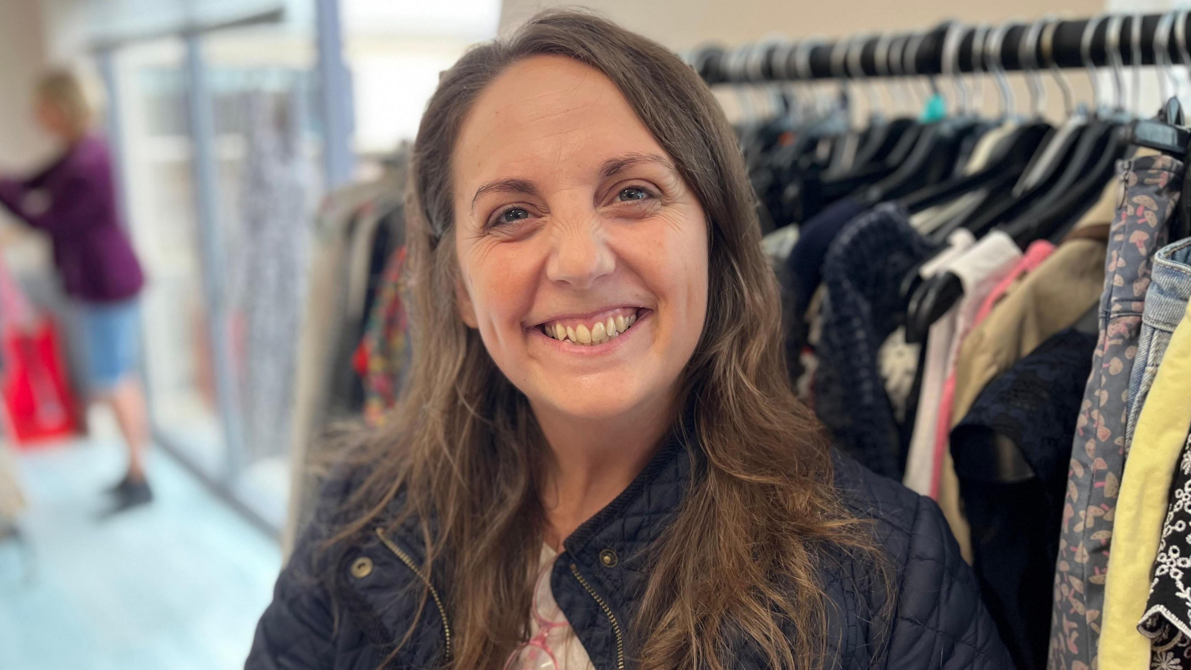 A woman with long brown hair smiling towards the camera. She's bearing her teeth and is standing in front of a clothes rack.