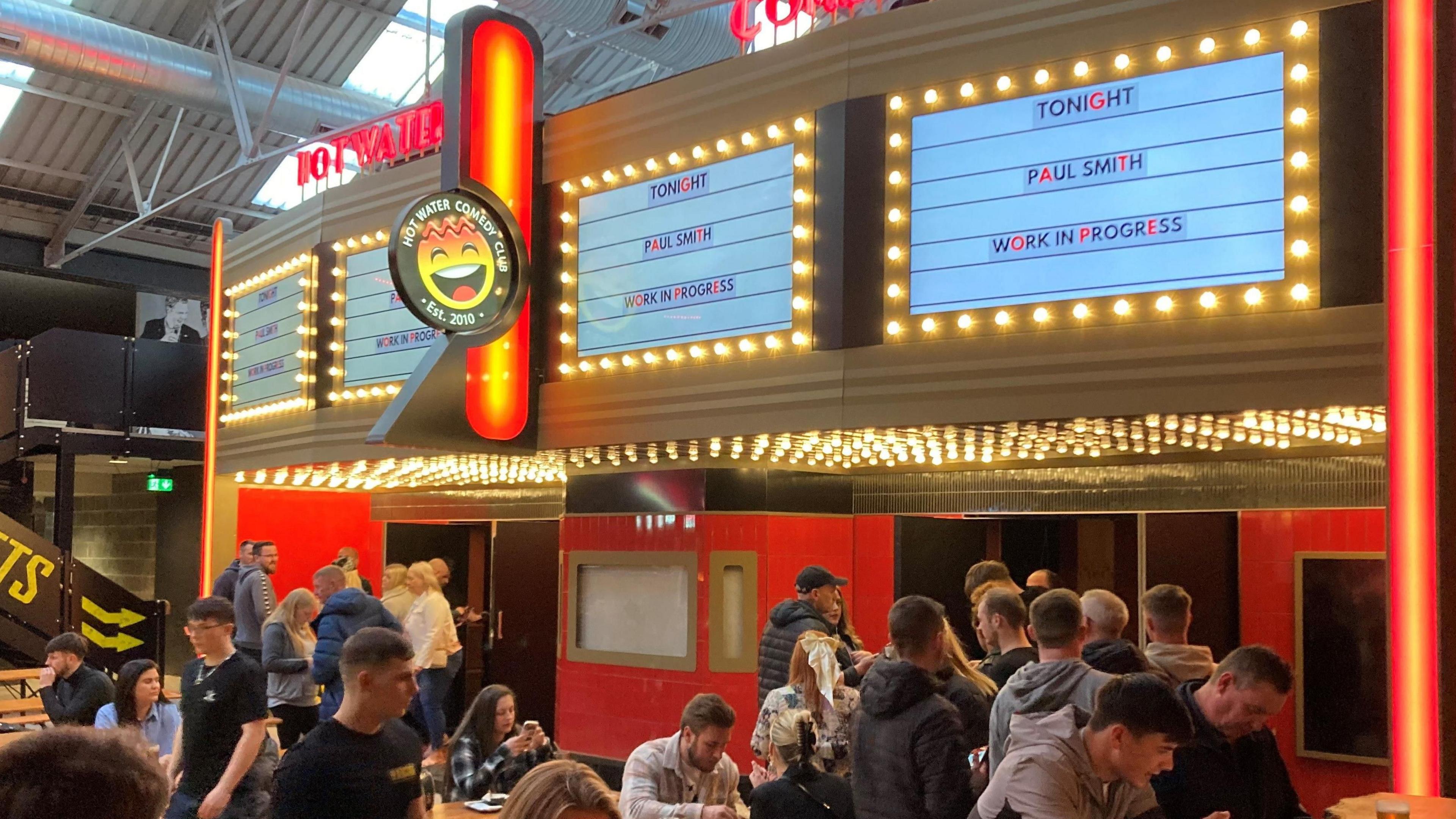 A crowd of people queue outside the entrance to the Hot Water Comedy Club venue at Blackstock Market, which has signs saying 'Tonight Paul Smith'. 