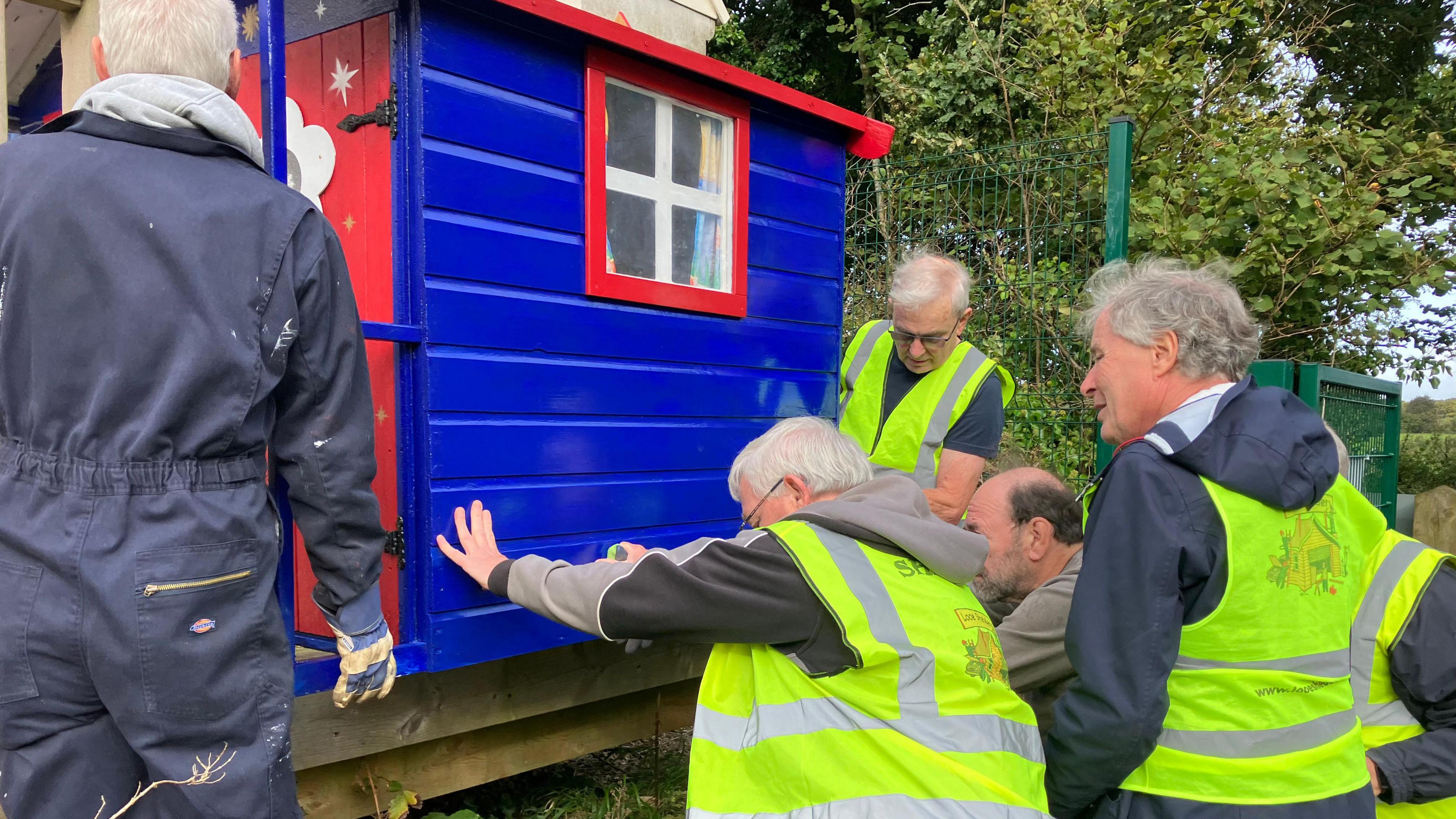 Several older men in high visibility yellow jackets pushing a Wendy house waiting into place.