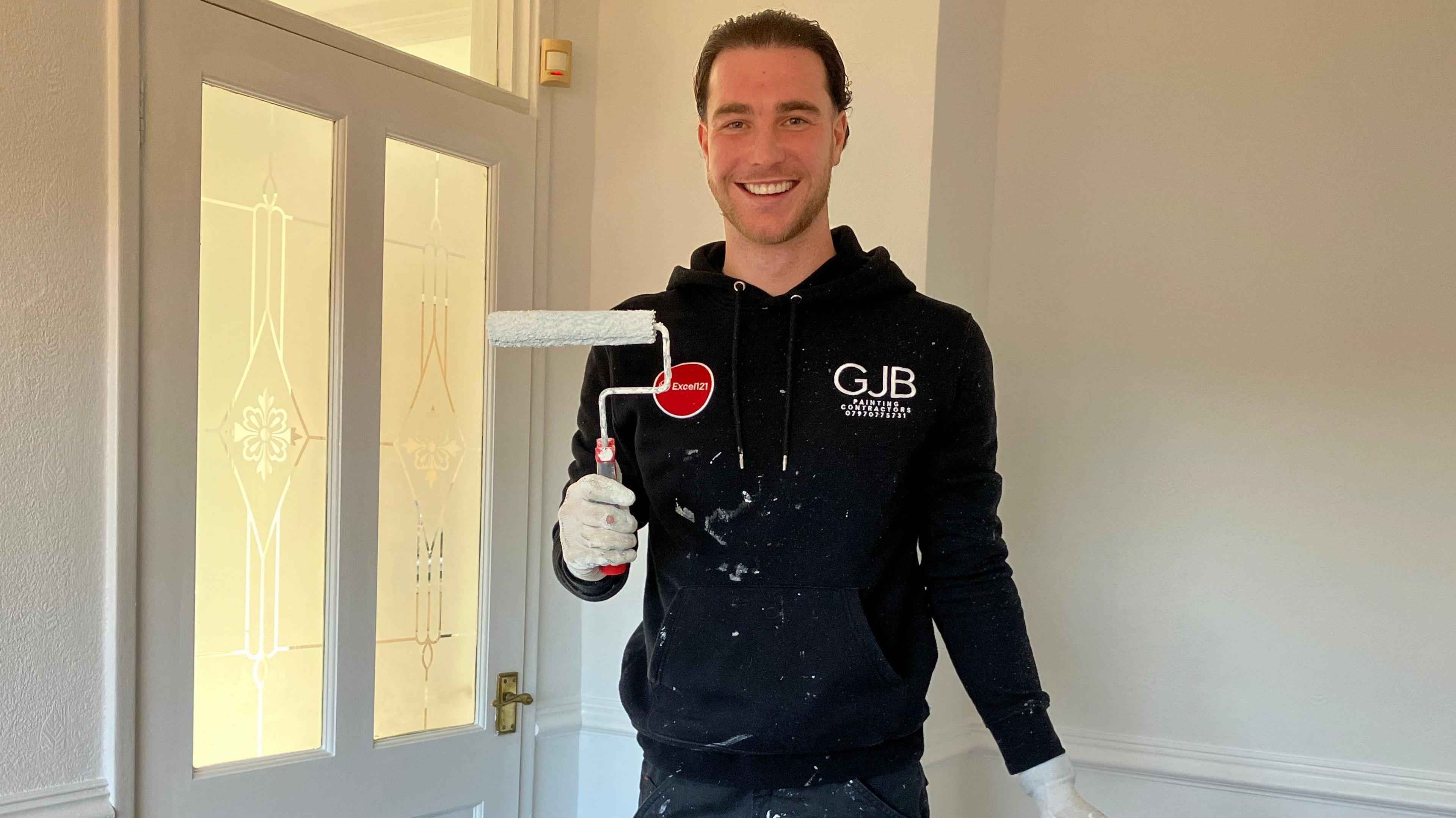 Louis Britton, who has short brown hair, stands inside the hallway of a house holding a paint roller. He is smiling at the camera and is wearing a black hoodie which has splotches of white paint on it. 