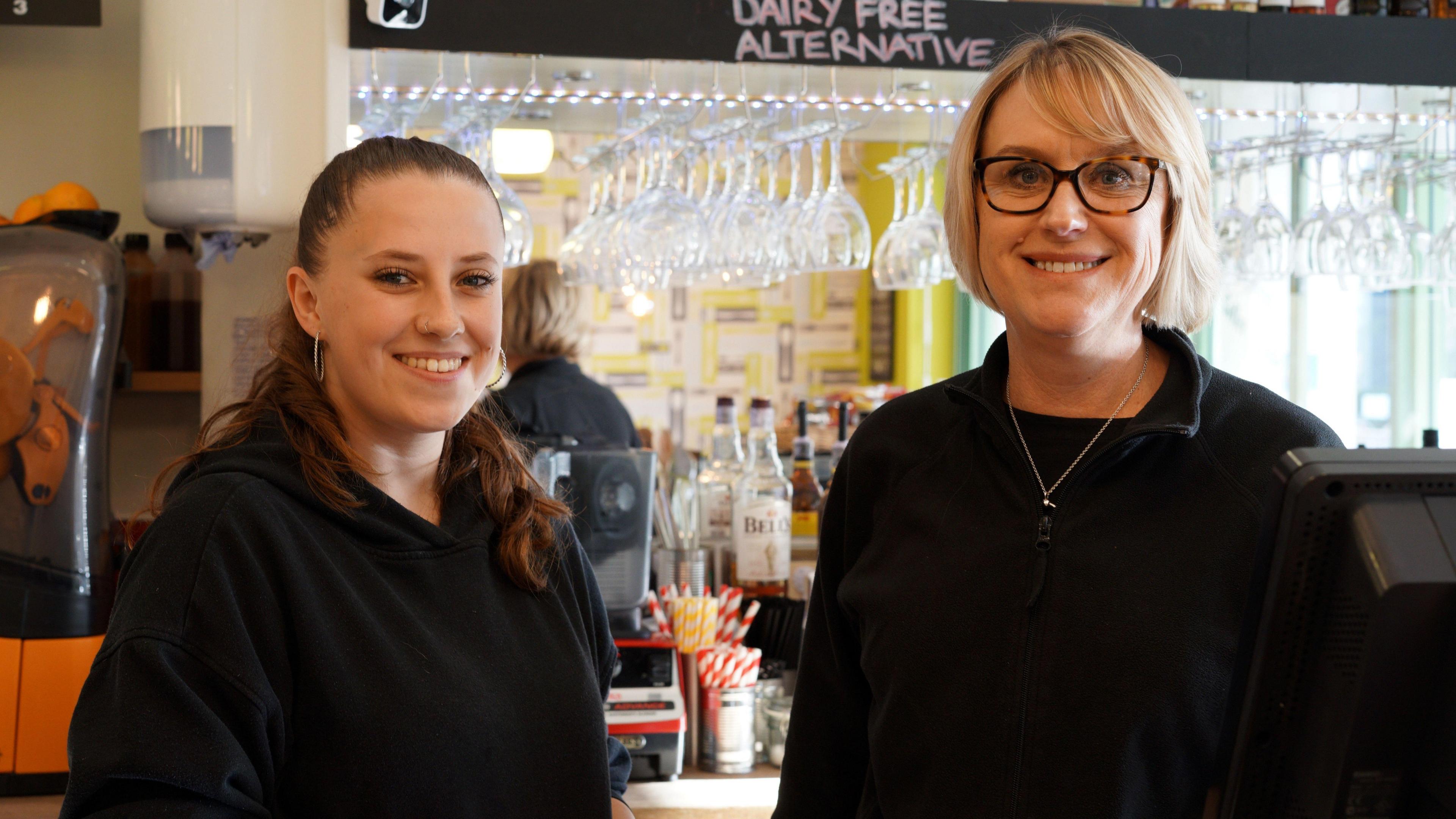 Christina and Claire are both wearing black tops and standing behind the coffee shop counter, smiling. In the background is a mirror and hanging up wine glasses.