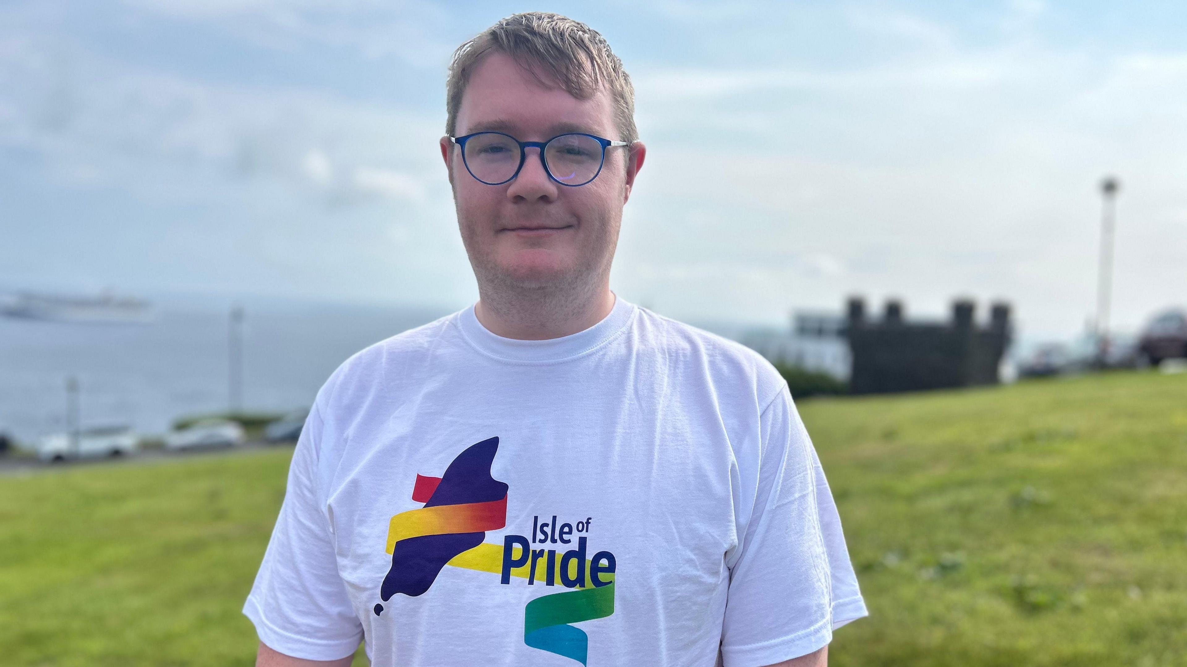 James Cherry stands on a grassy hill, he wears blue spectacles and a white t-shirt with the Isle of Pride logo. It is colourful, with a rainbow ribbon that floats around the name of the charity and a purple Isle of Man-shaped motif. 