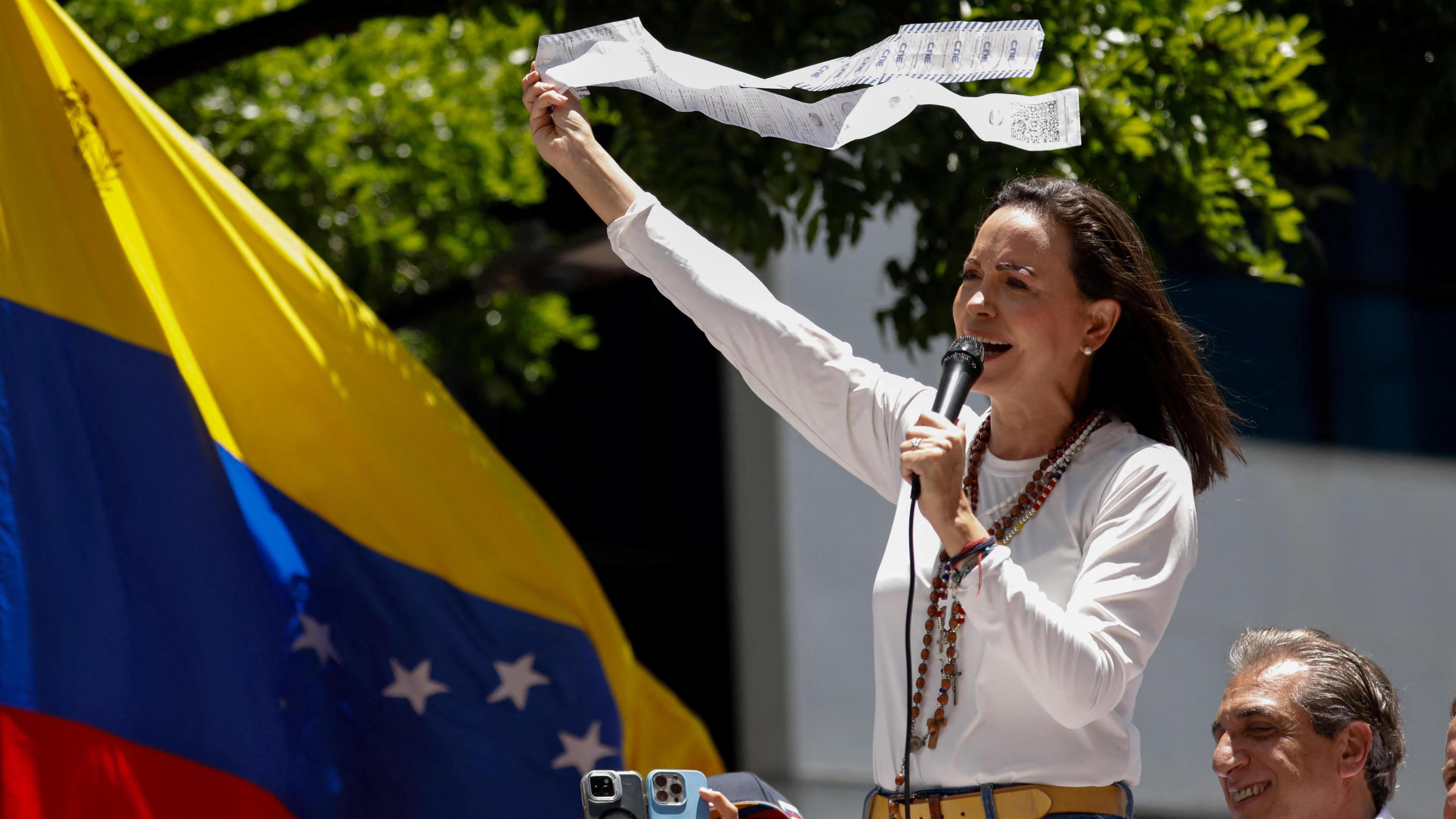 Venezuelan opposition leader María Corina Machado waves a voting tally in front of a Venezuelan flag as she speaks during a rally.