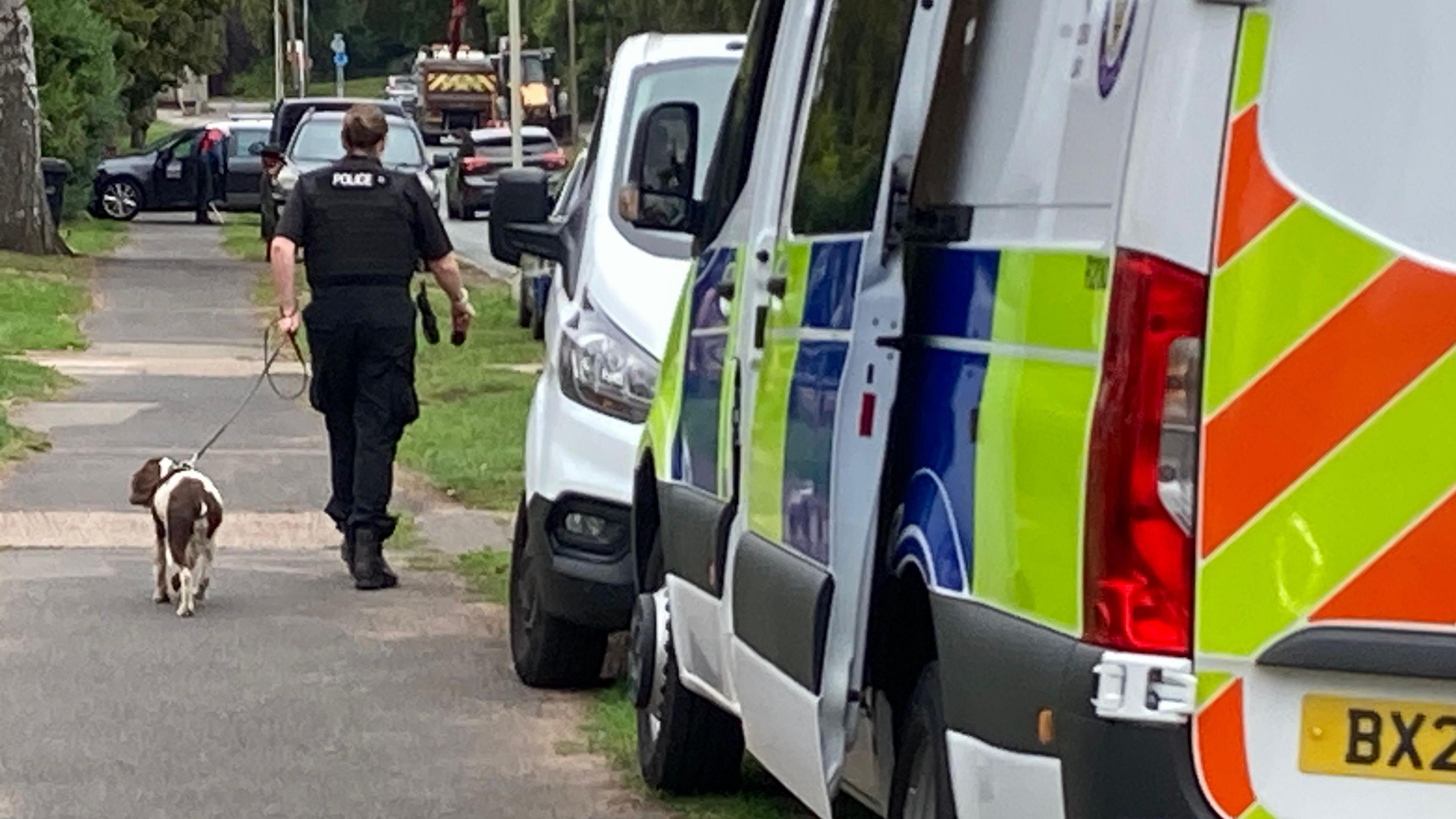 A police officer walks along a pavement with a dog on a lead next to several emergency service vehicles