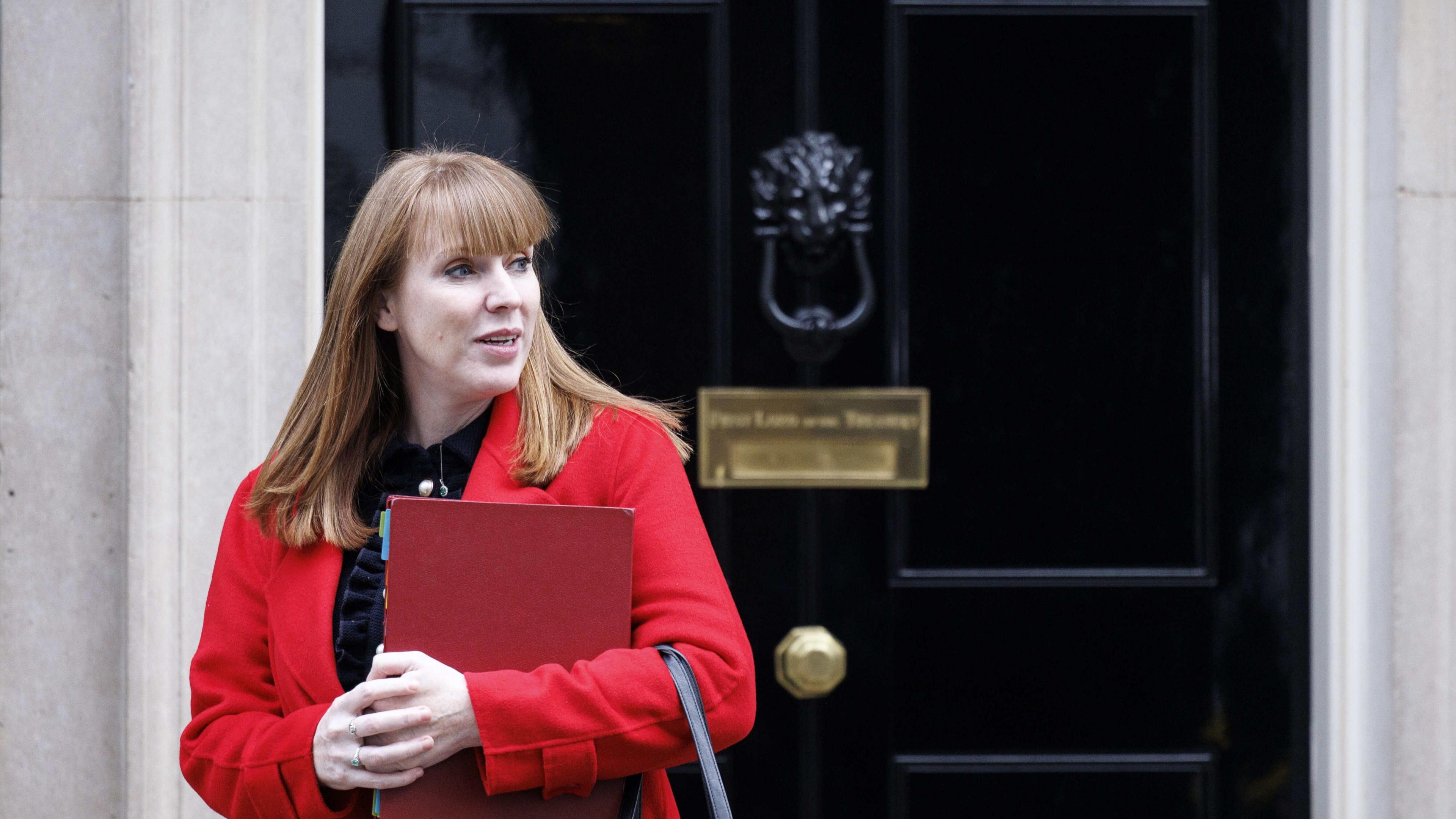 Deputy Prime Minister Angela Rayner wearing a black top and a red coat and holding a file outside 10 Downing Street