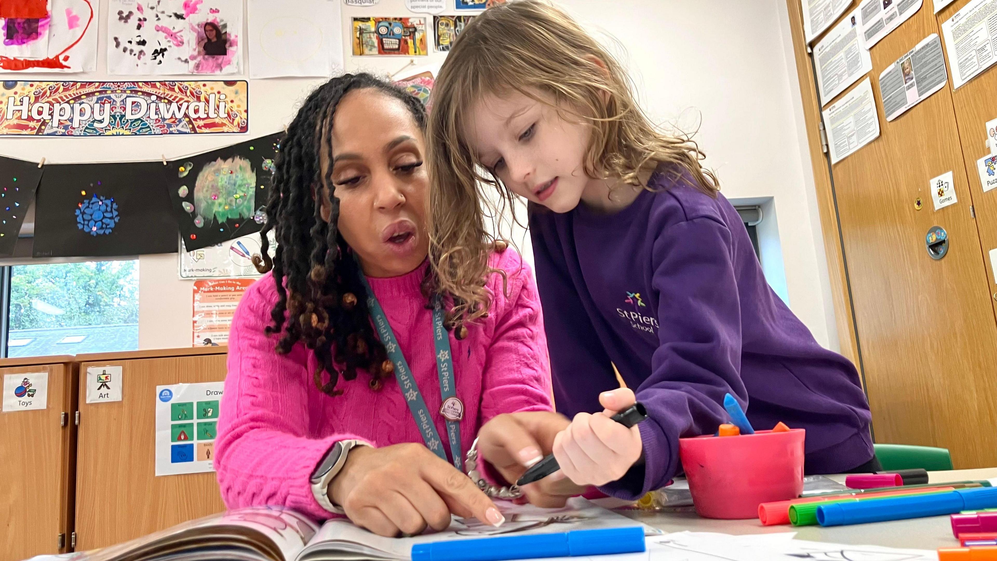 A teacher from St Piers School sitting at a classroom table desk pointing and showing a young child a sentence in a reading book while the child holds a pen and there are coloured pencils and pens on the table.