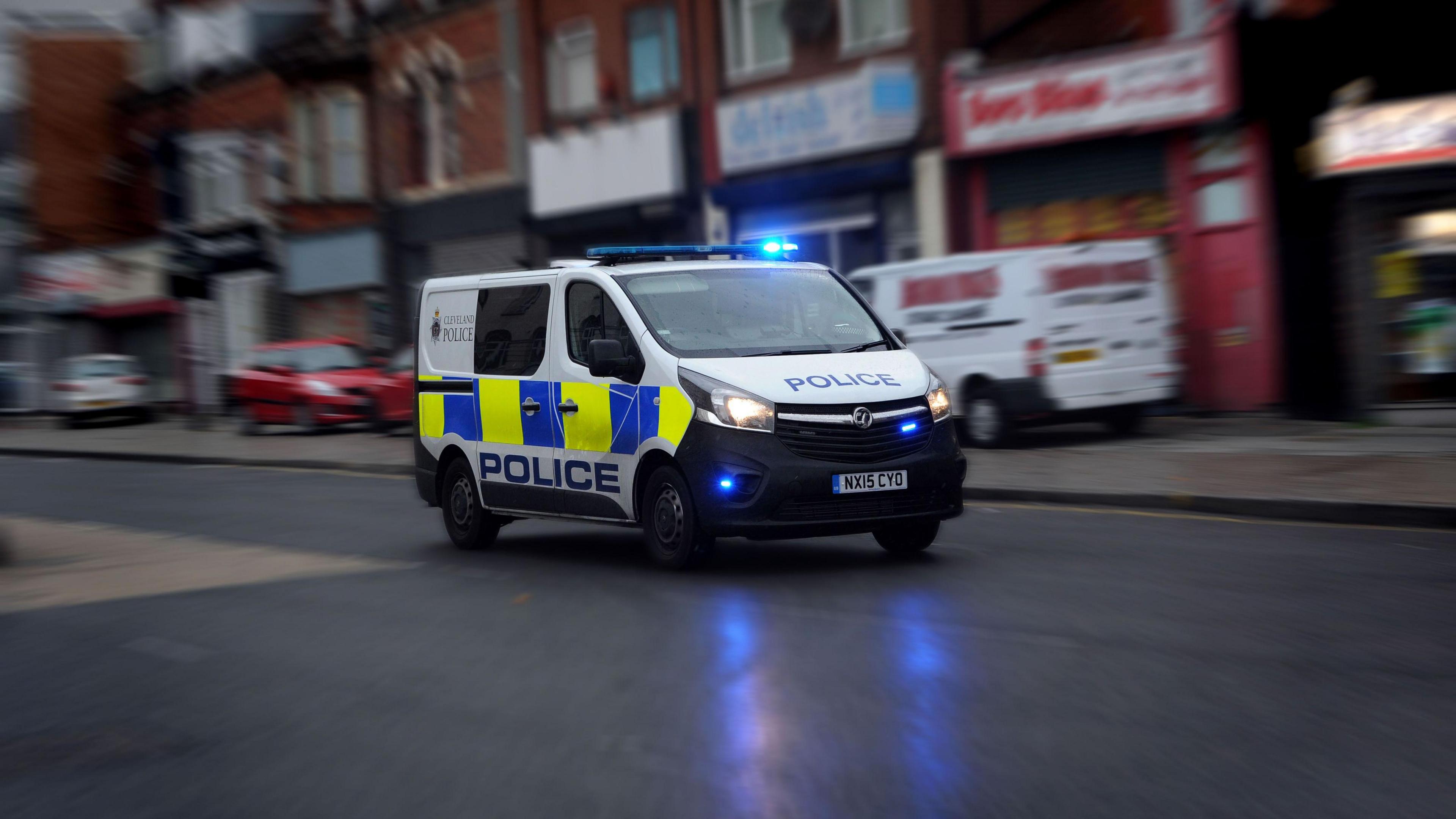 A Cleveland Police van driving through a high street. The vehicle's blue lights are flashing.