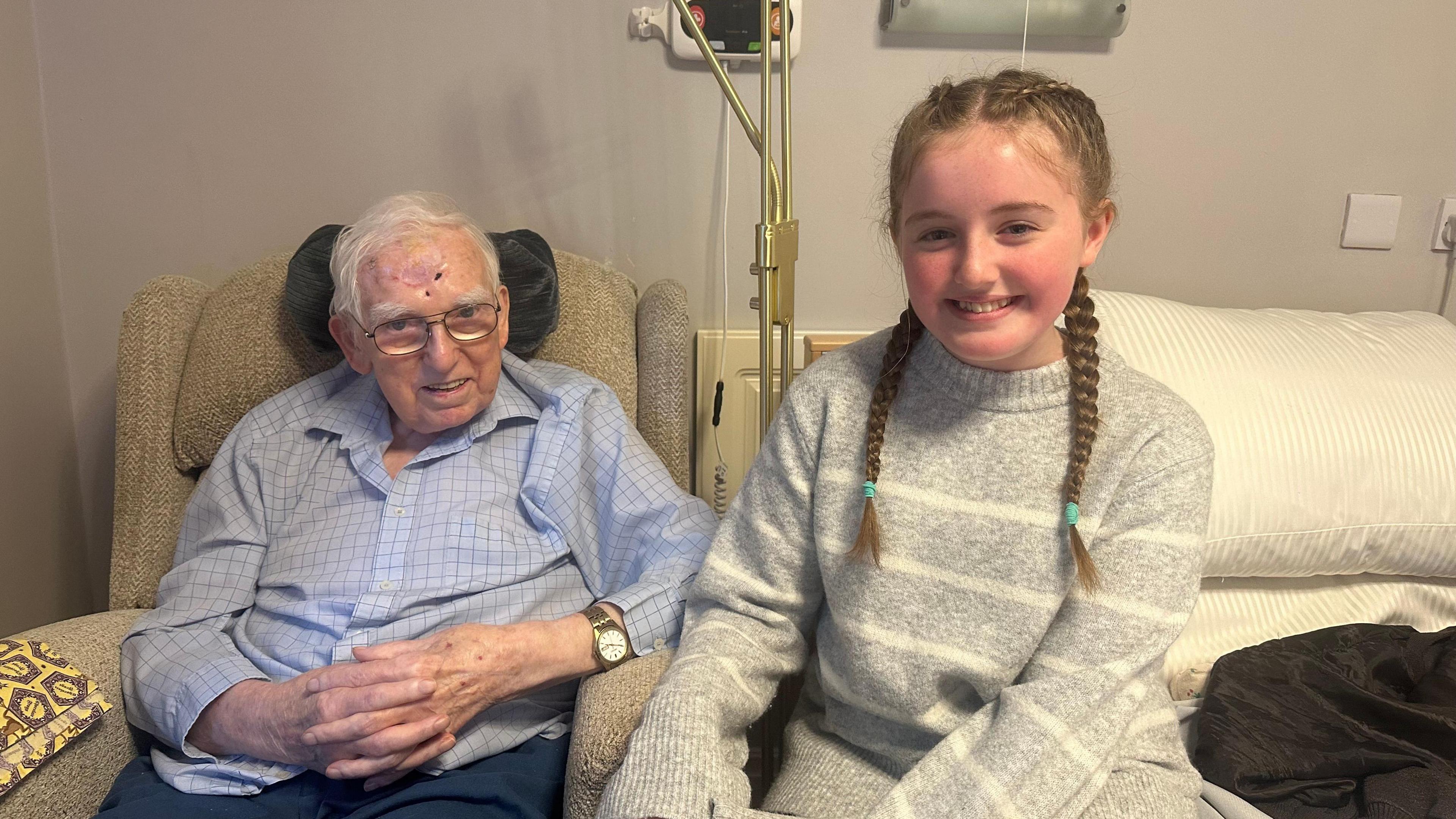 A man in his nineties sits in a care home chair. He has white hair and glasses and wears a blue shirt. A brown watch is on his right wrist. Next to him is an 11 year old girl. She has mousey brown hair tied in plaits and wears a grey jumper. They smile.