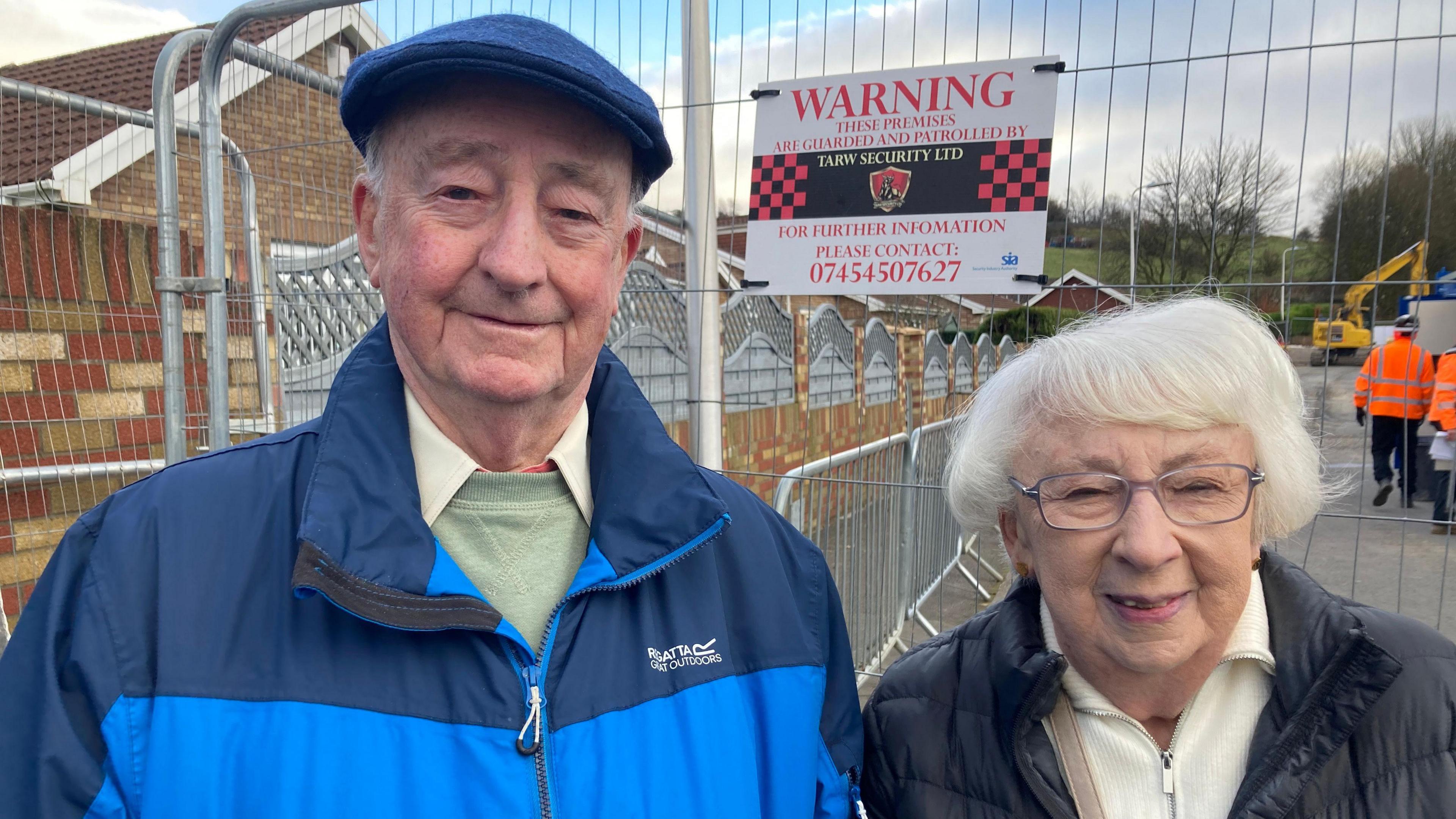 Meurig and Sheila Price smile looking at the camera standing on their street. There is a metal barricade behind them which borders the sinkhole making it clear that they are currently unable to return home.