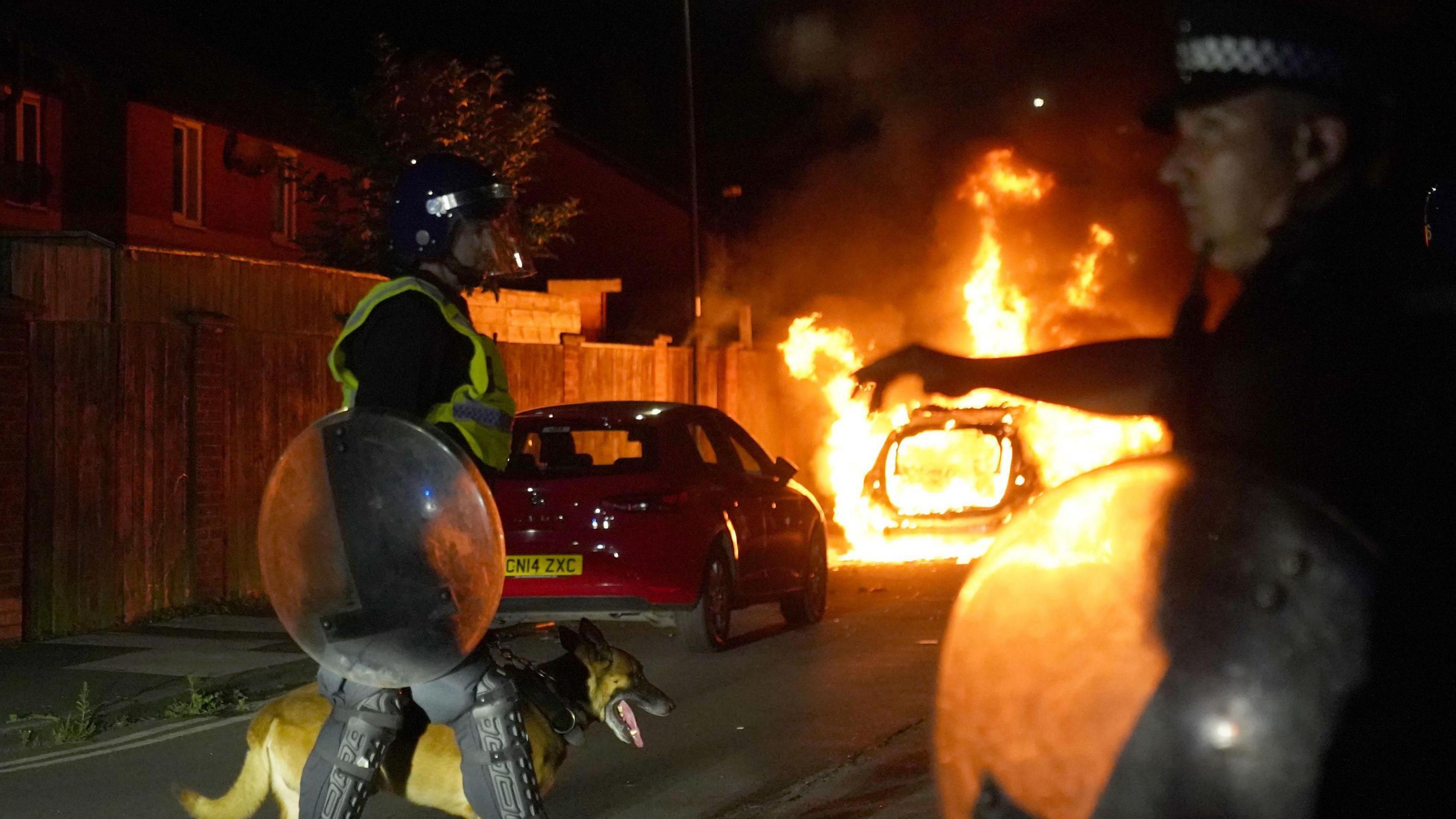 A night scene from a street in Hartlepool. Two police officers in riot gear, one with a police dog, stand side on and facing in opposite directions in front of two cars, the second of which is on fire.