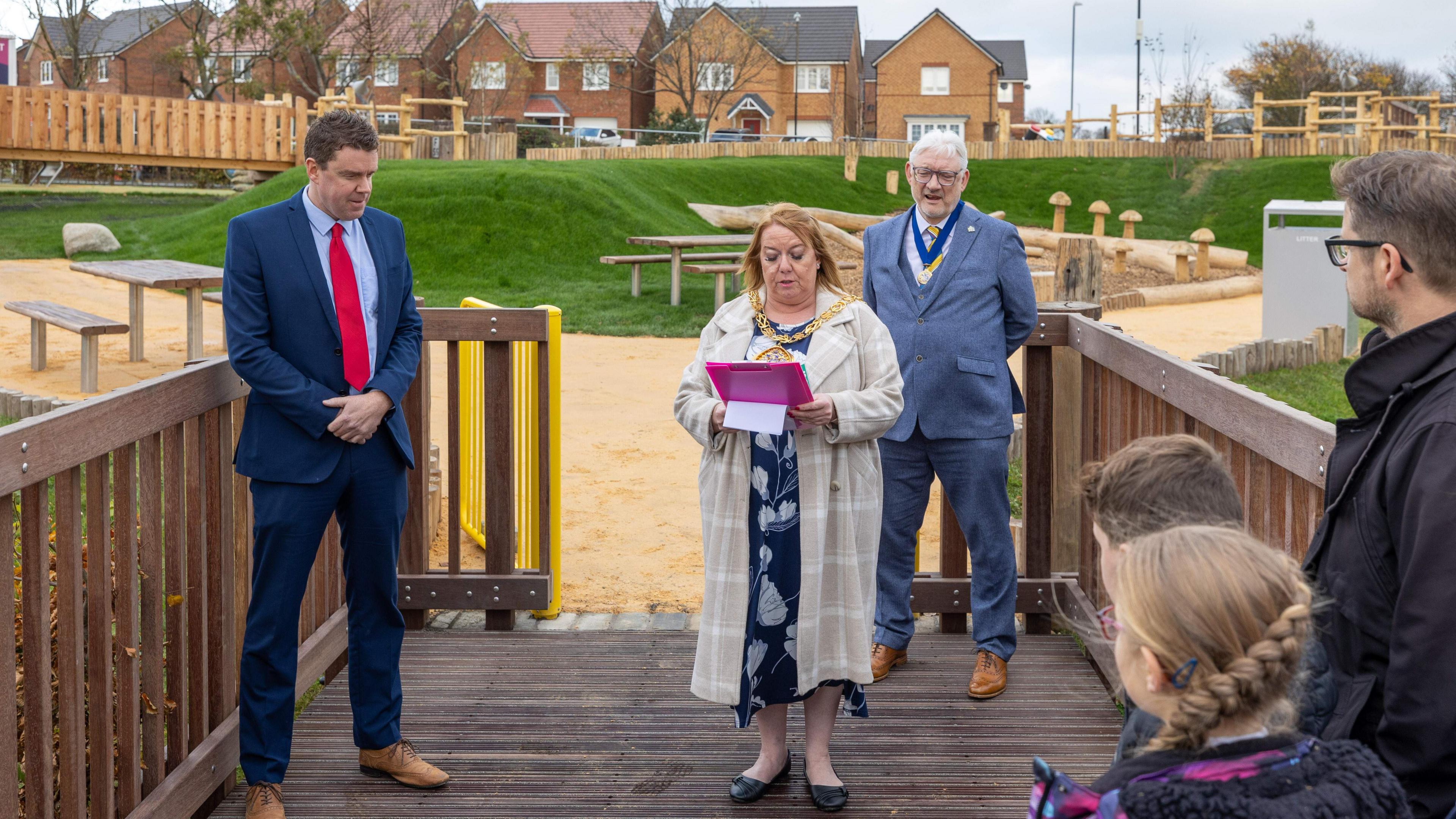 Mayor of Sunderland councillor Allison Chisnall is holding a clipboard and reading a speech as she opens the play park. Children and other officials can be seen listening to her.