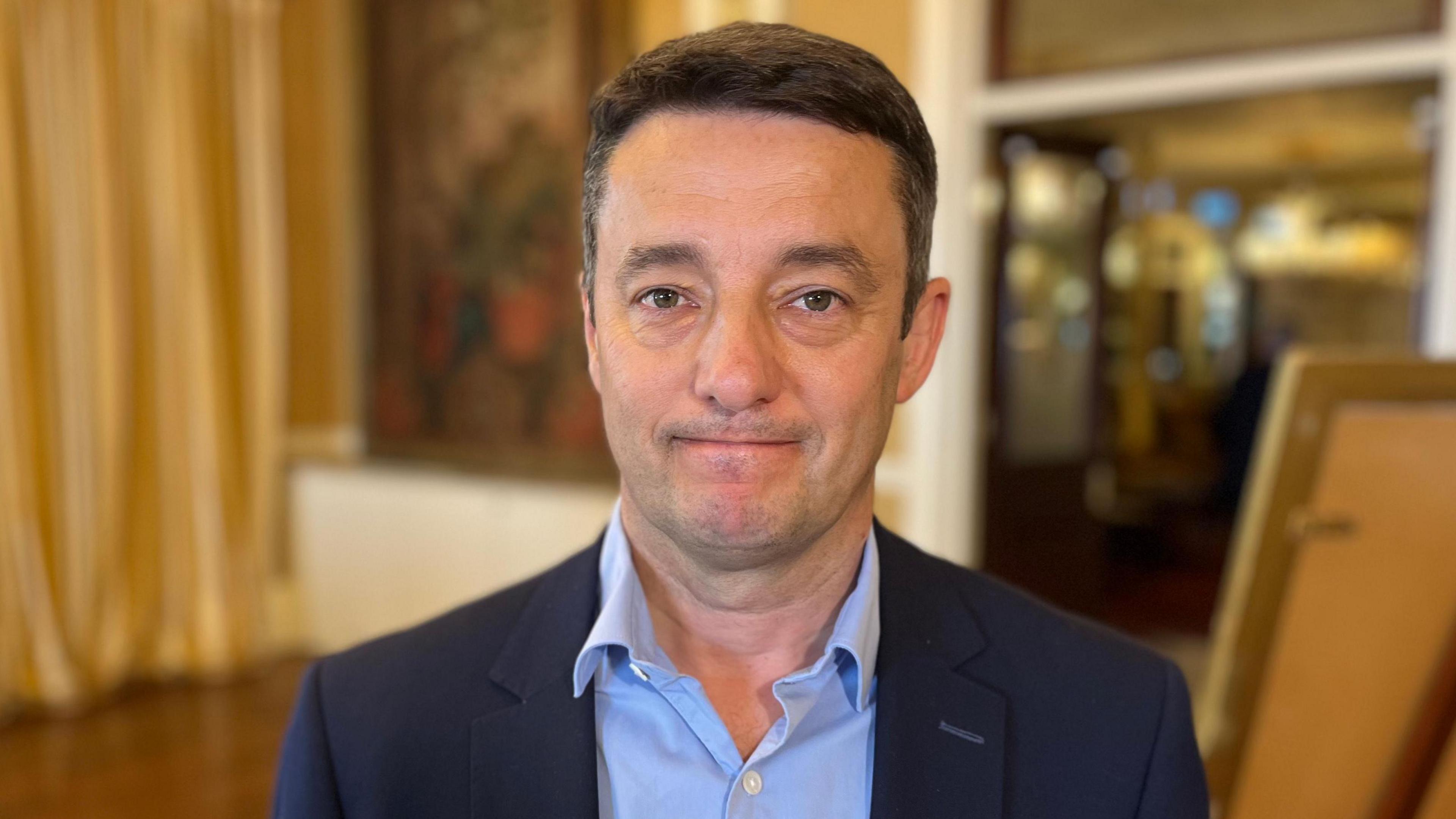 Boley Smillie - a man wearing a navy suit and light blue shirt, standing in a large hall room which is blurred.