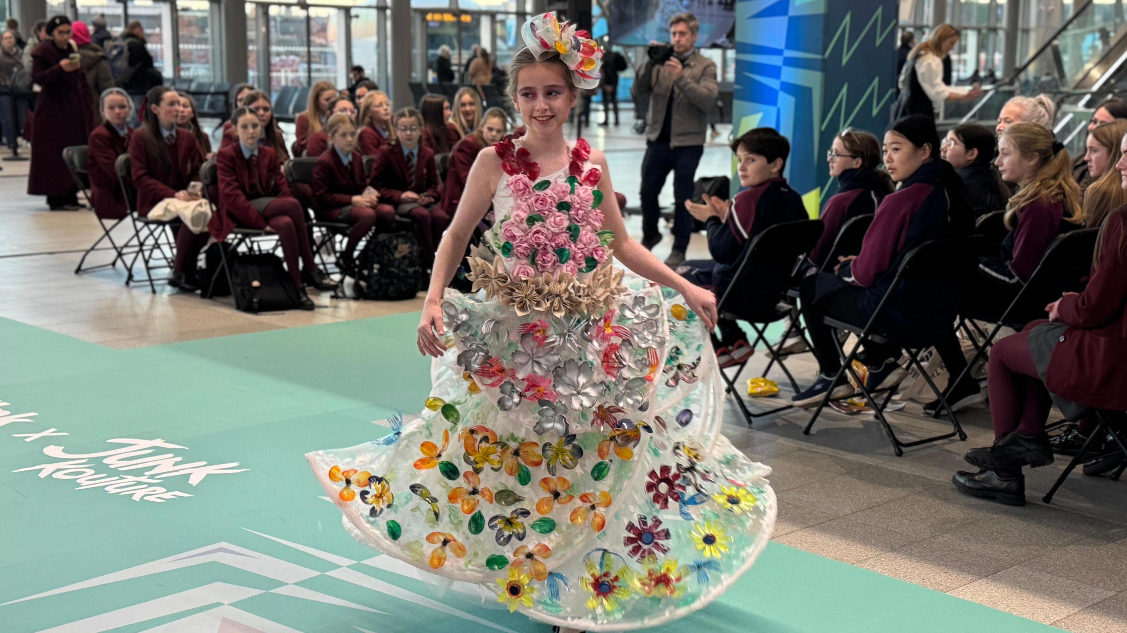 A student wearing a dress made of plastic with different coloured flowers along the dress and head piece. 