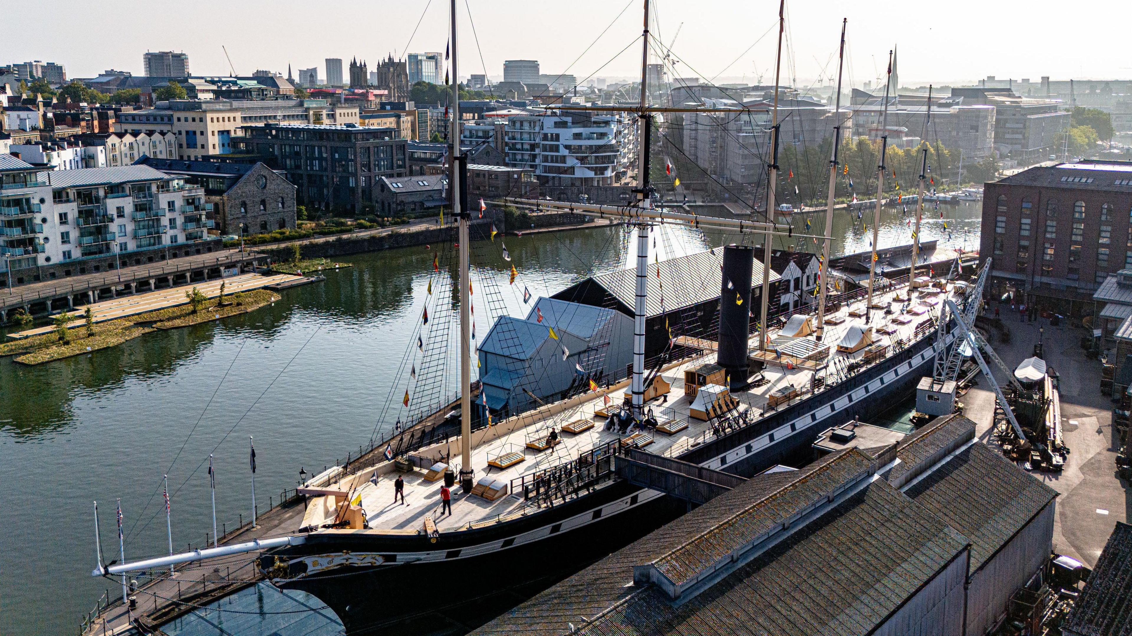 An aerial view of the SS Great Britain ship which is docked on Bristol's Harbourside. The ship is surrounded by apartments and tall buildings. The photo has been taken on a cloudy but clear day.
