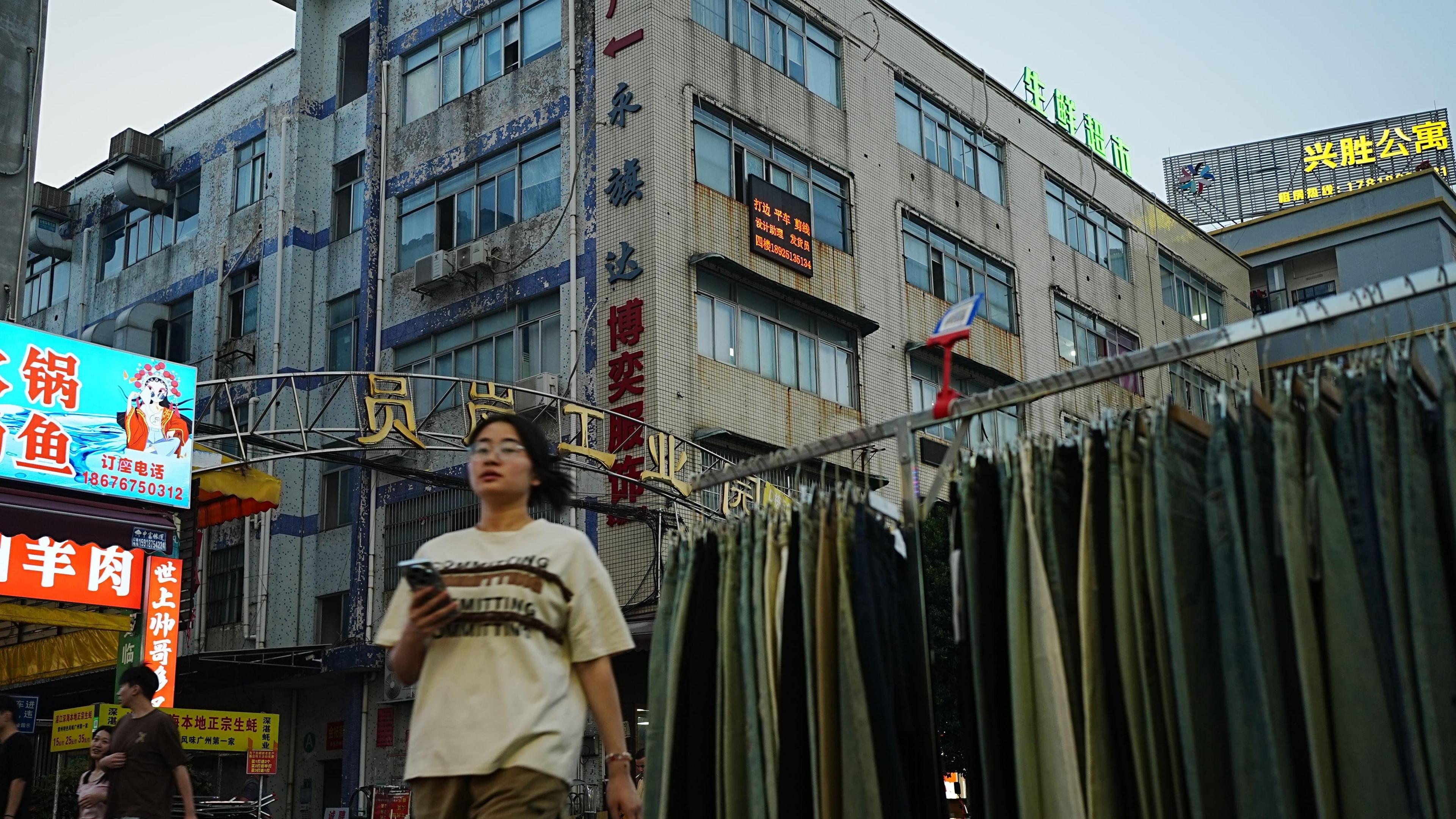 A women wearing a t-shirt and shorts walks past a row of jeans on display on the street in the late evening. Behind her are buildings which house 
the factories.