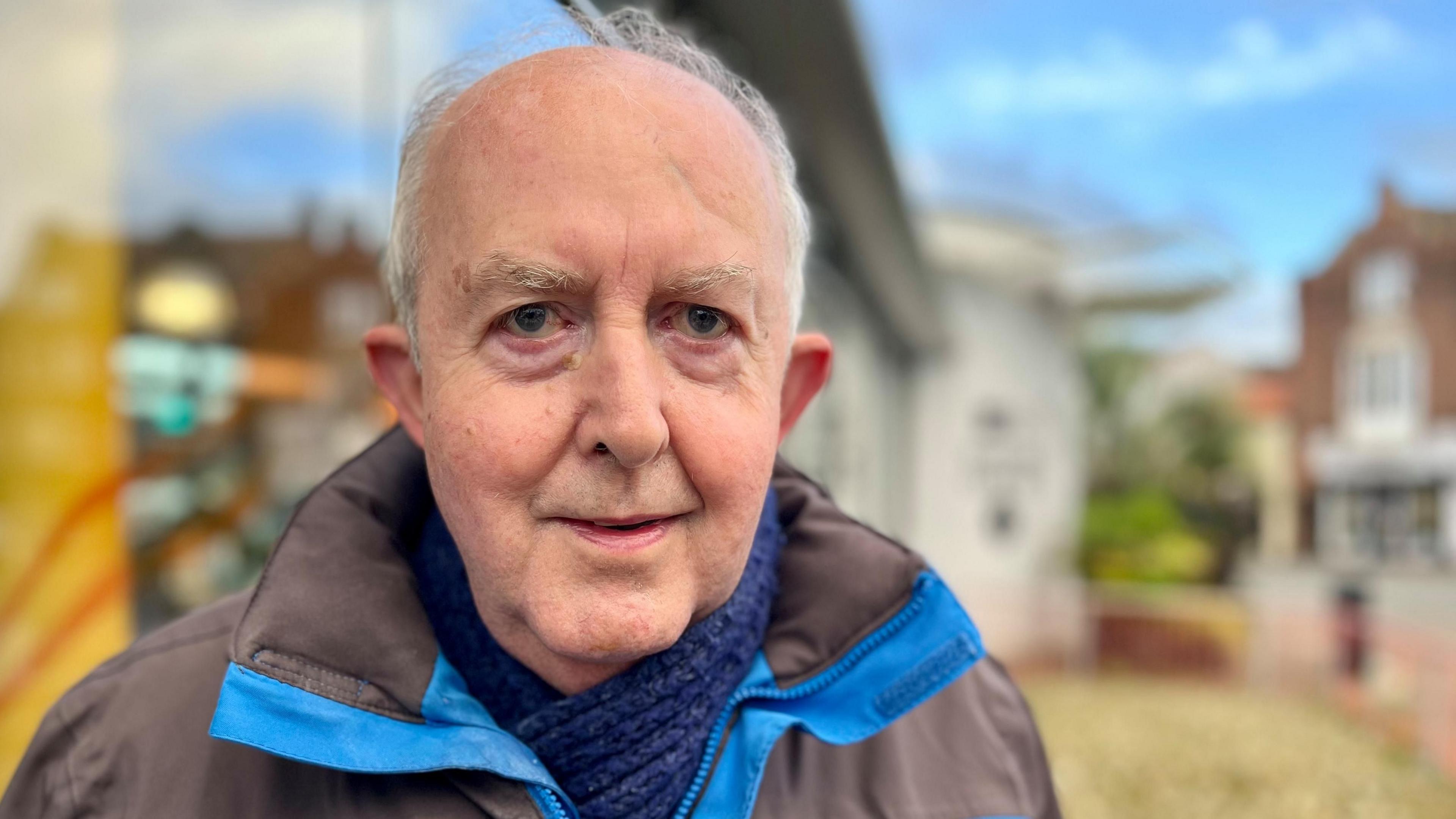 Trevor Hatter is looking into the camera with the visitor centre in Cromer behind him. He is wearing a black and blue jacket with a blue scarf.