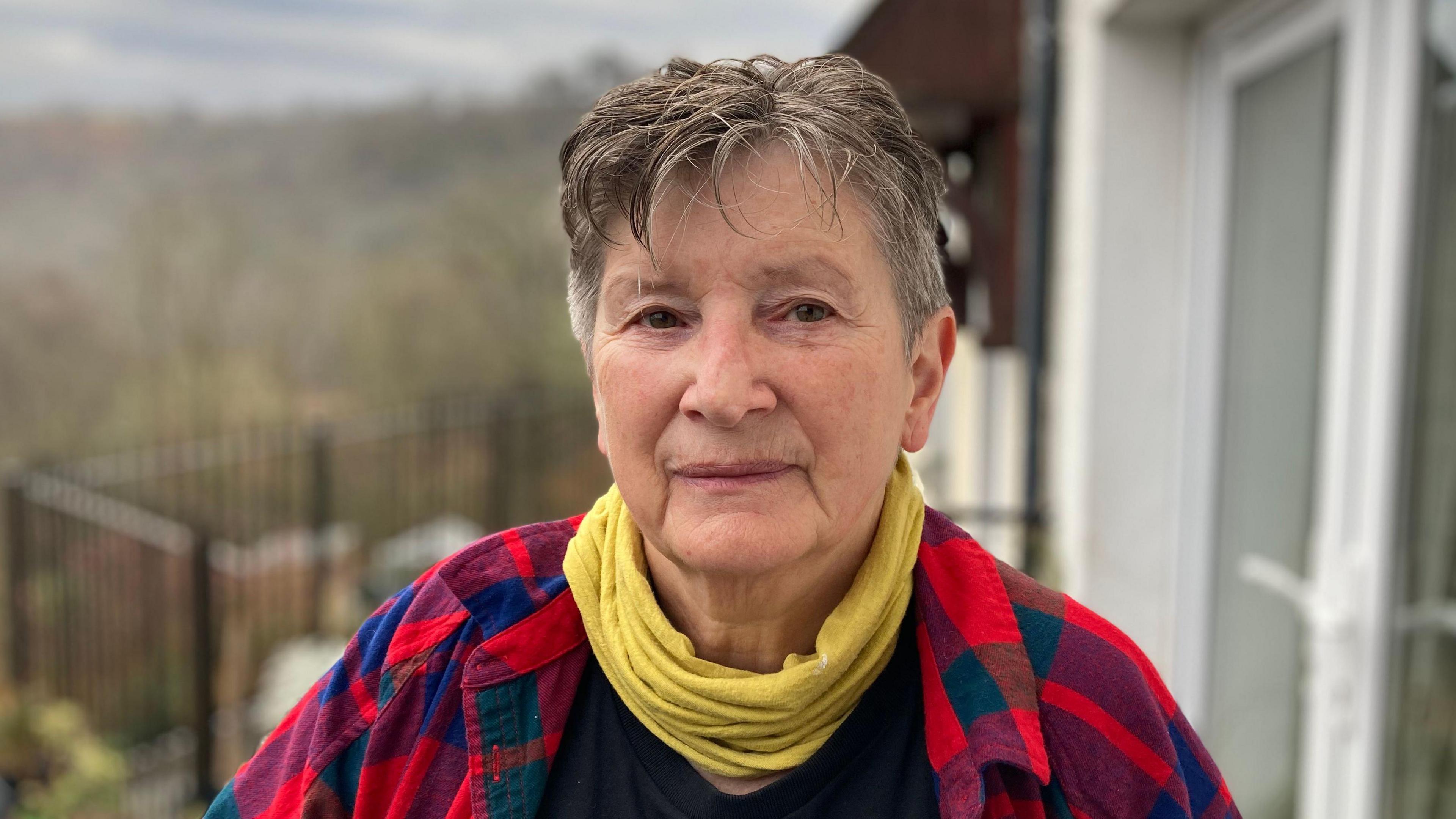 A woman with short grey and brown hair, wearing a yellow snood and a red and blue plaid jacket