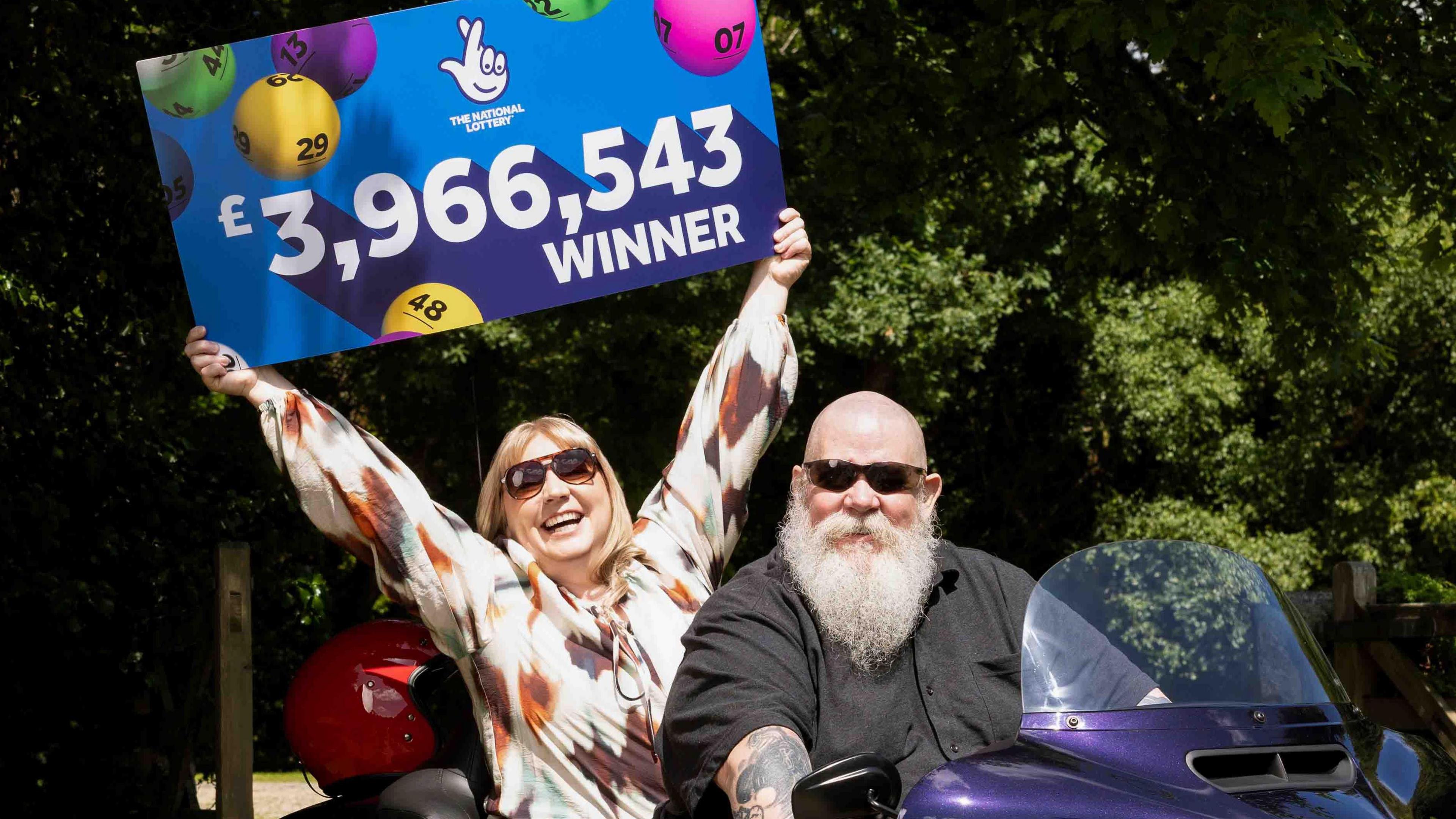 A woman holding a sign that says "The National Lottery and £3,966,543 winner". The background of the sign in clue and there multicoloured bingo balls. There is a man sat in front of the woman on a purple Harley-Davidson trike bike. There are trees behind them and a wooden fence.