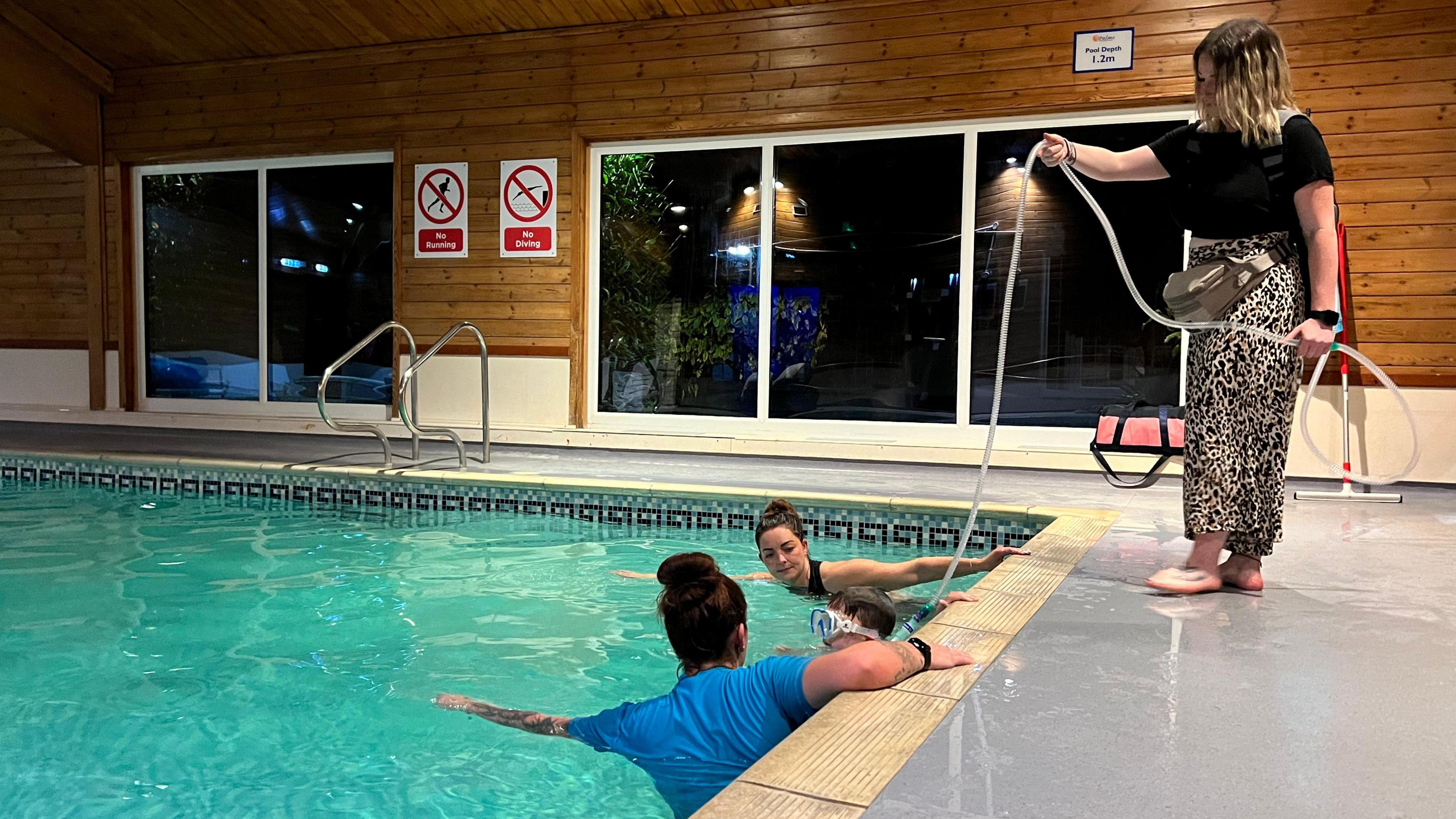 Henry, who is wearing goggles, prepares to push off from the edge of the pool with Kayleigh and Shevonne either side of him. The carer stands behind them at the side of the pool and holds the tube connecting the ventilator to Henry's neck. 