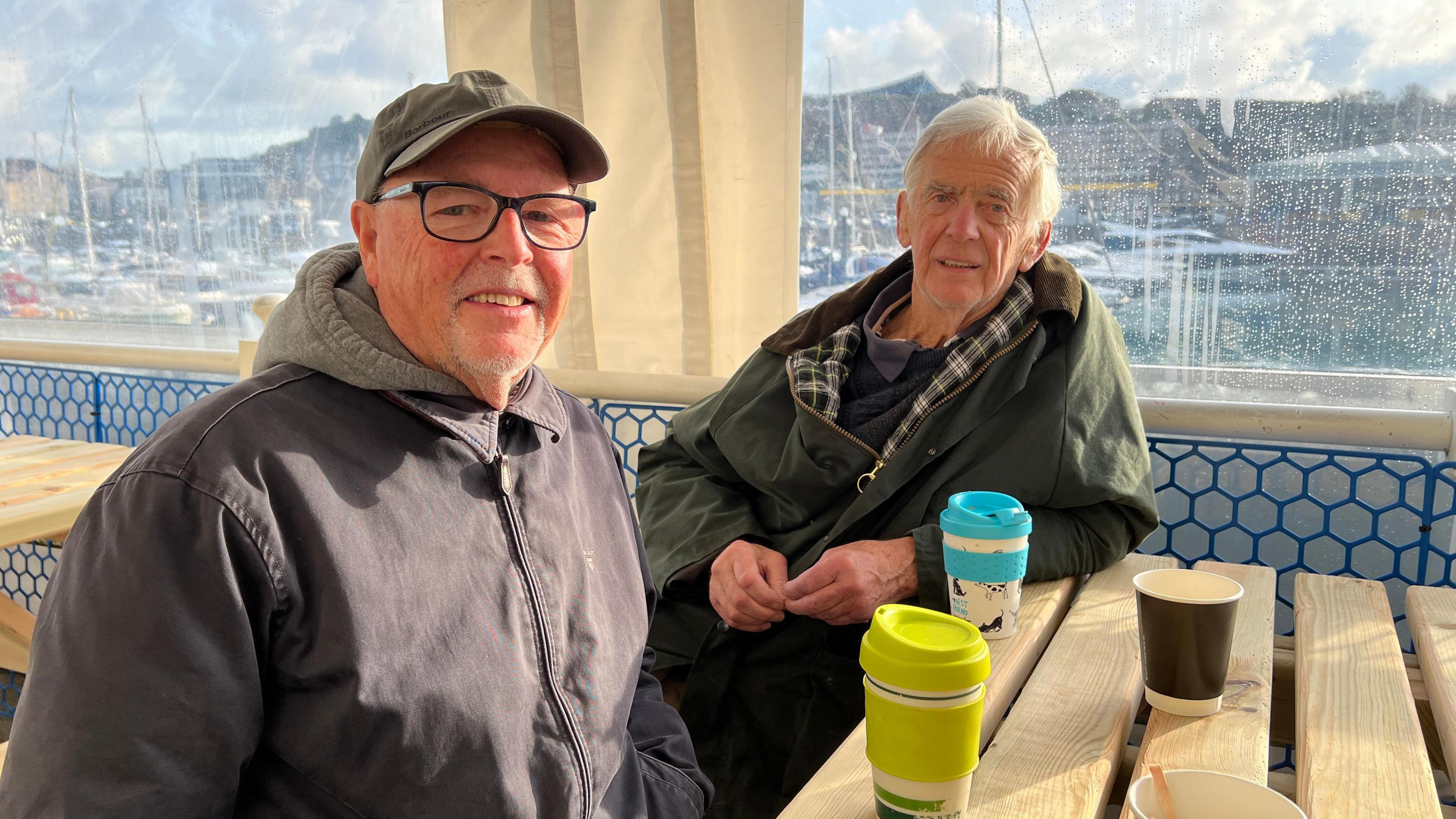 Two old men with grey hair sit under a plastic cover on wooden tables drinking out of reusable cups.

The man in the foreground has a zipped-up blue jacket, glasses and a green cap while the man behind leans on the table in a green wax jacket.