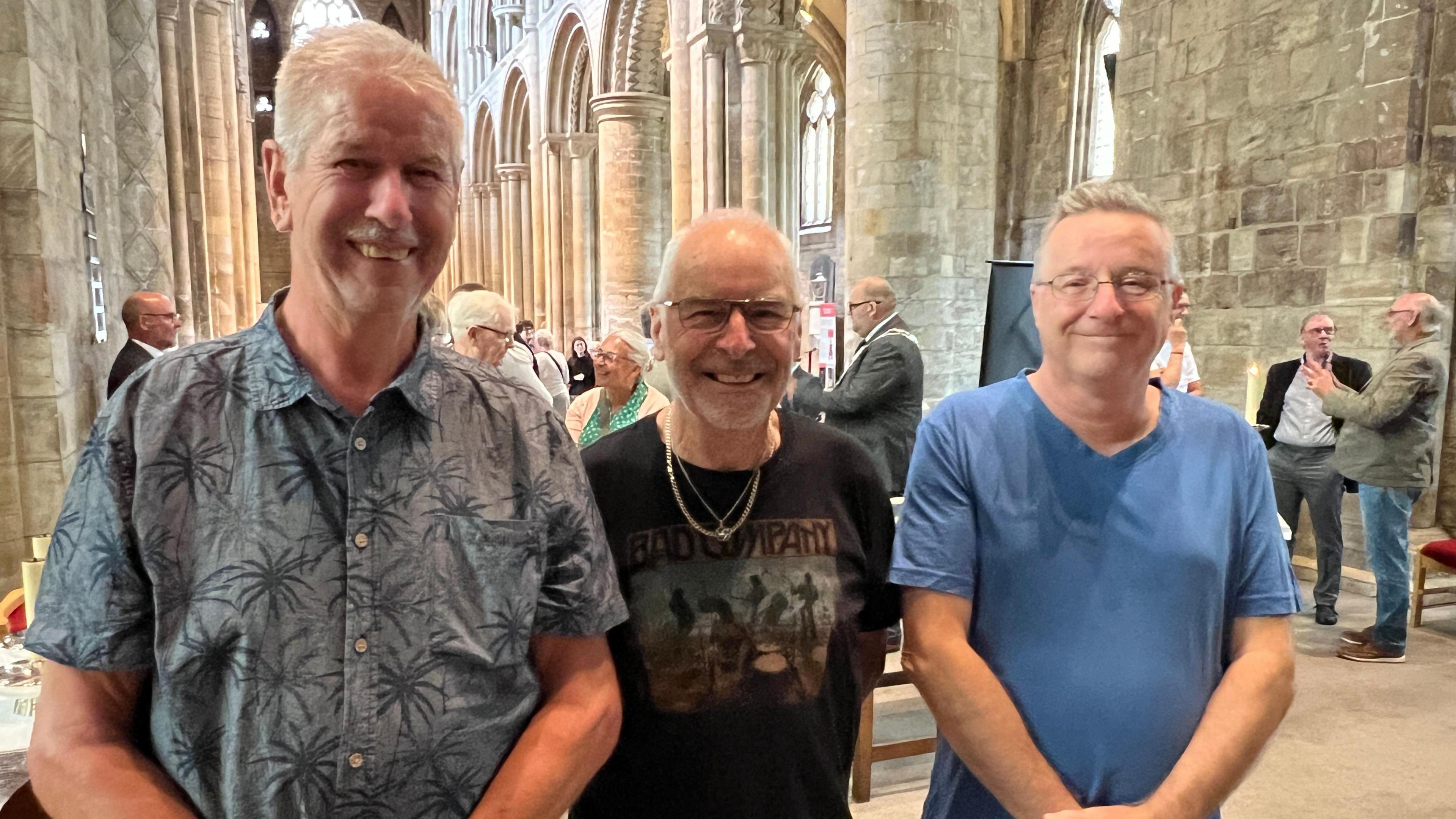 Former shipbuilders Phil Moat, David McMillan and Paul Welch are standing in a line on the altar at Selby Abbey. Phil is wearing a shirt with palm trees on it, David is wearing a black T-shirt and two gold necklaces and Paul is wearing a blue T-shirt. They are all smiling.