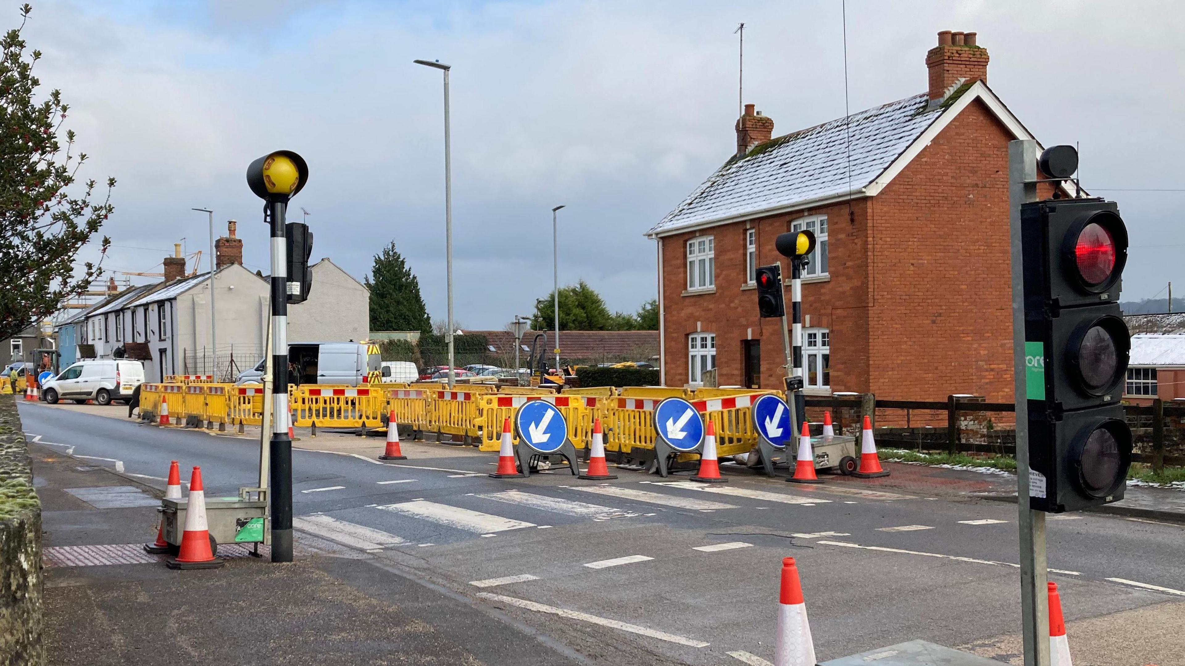 Roadworks and temporary traffic lights in front of a zebra crossing. There's a house in the background with road work barriers and cones in the foreground.