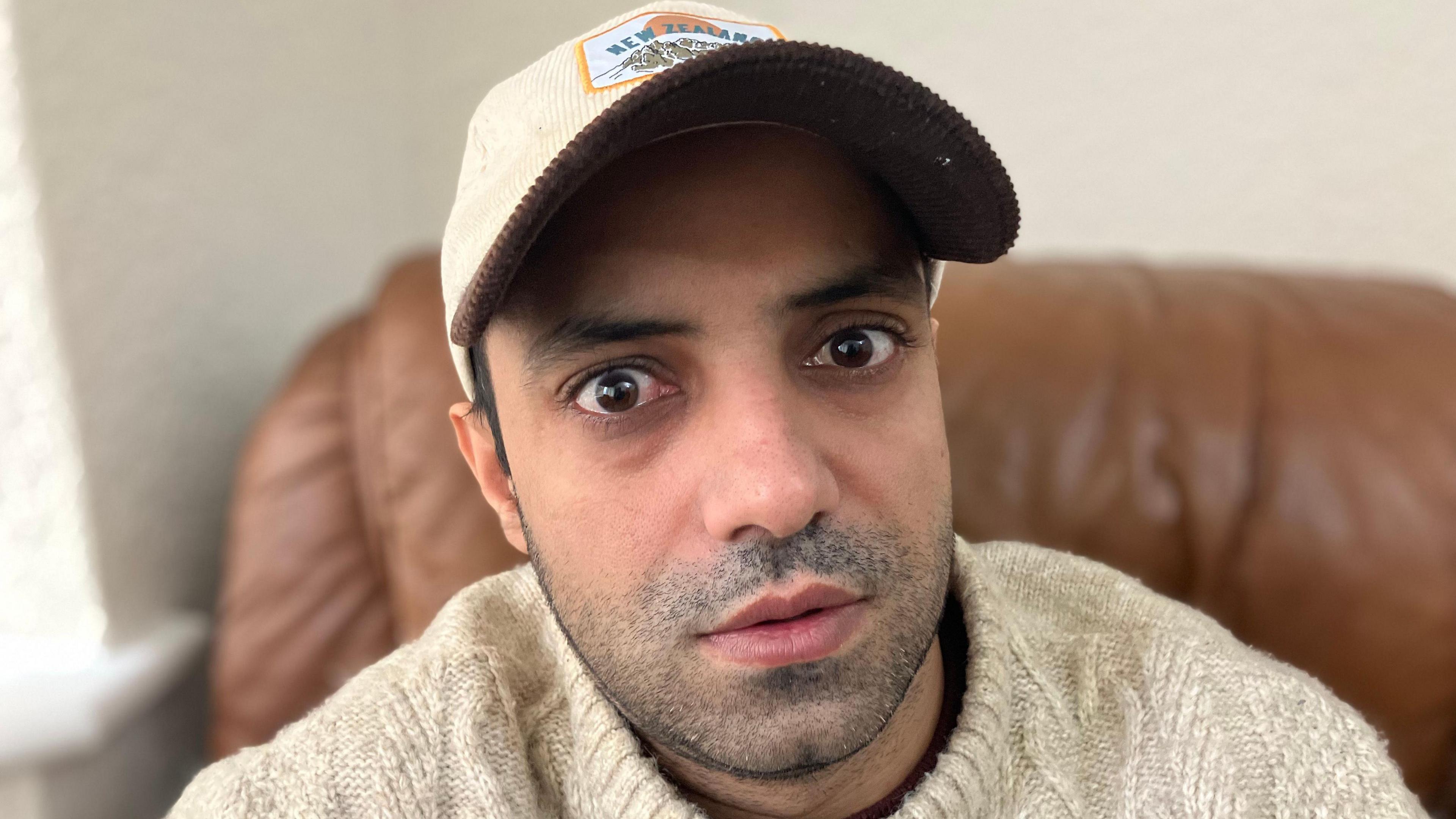 Nabeel Ahmad sat in a brown leather armchair, wearing a white woollen jumper and a white cap. He is unshaven and is looking at the camera.