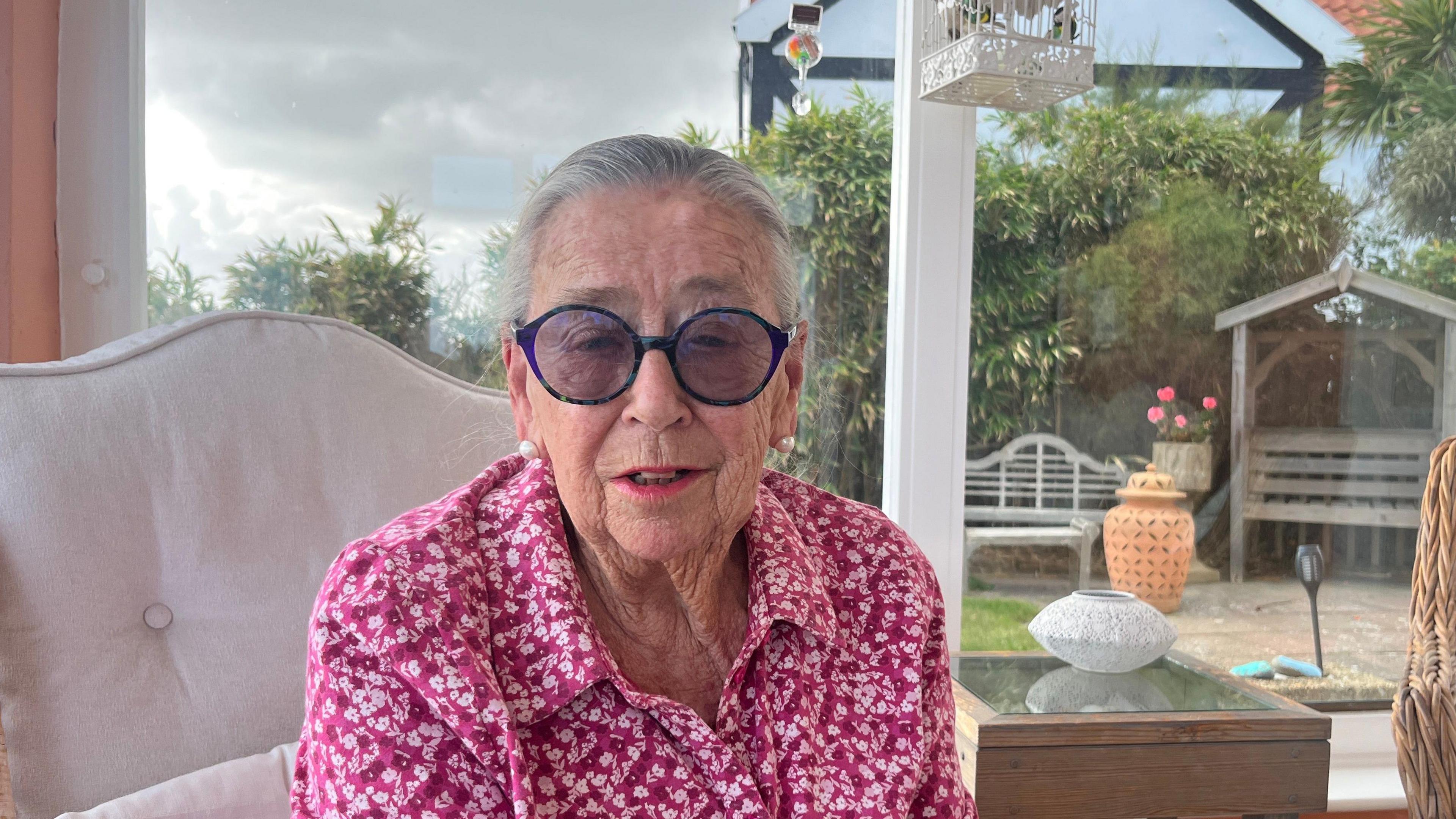 Shelley Cowlin wearing a pink top and blue glasses, she is say in a white chair in front of a window with a garden in the background. 