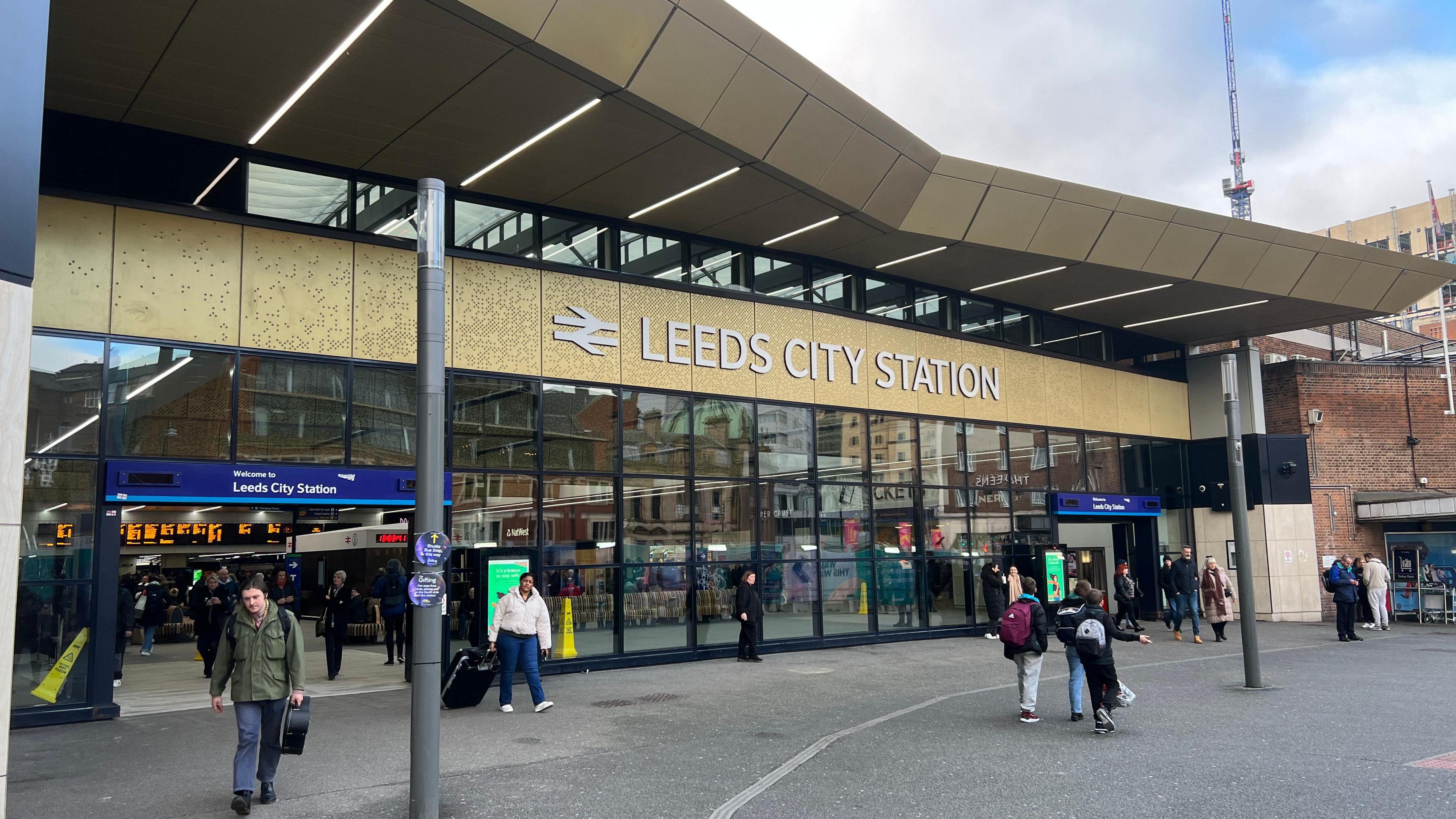 Outside Leeds City station. The building has a glass front, with the name of the station above the glass. It is a sunny day and people are walking in and out of the station.