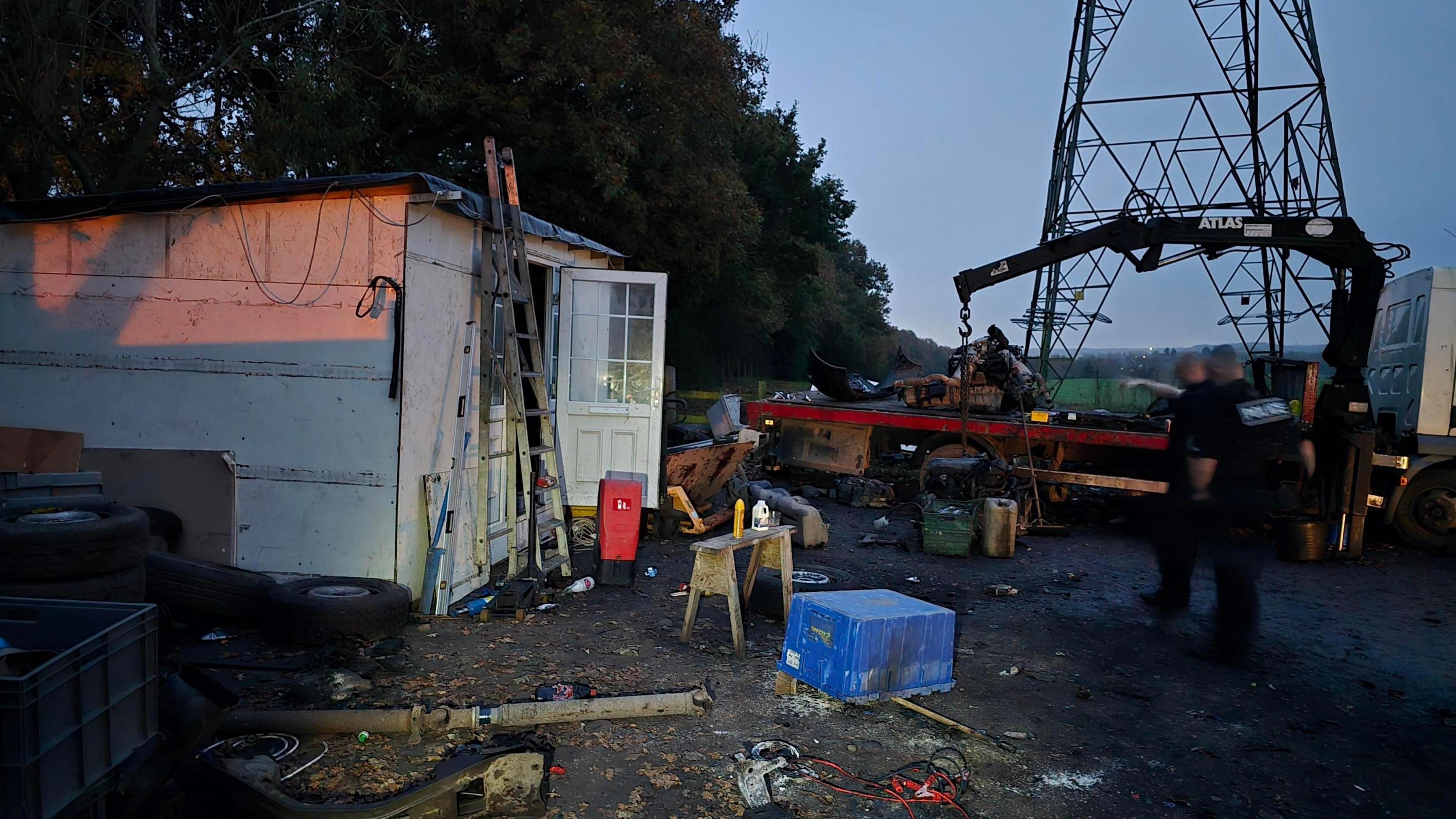 A suspected chop shop with work equipment on the ground, a small white hut with two ladders resting against it. A truck can be seen in the background and two police officers wearing black uniforms are looking at the site.