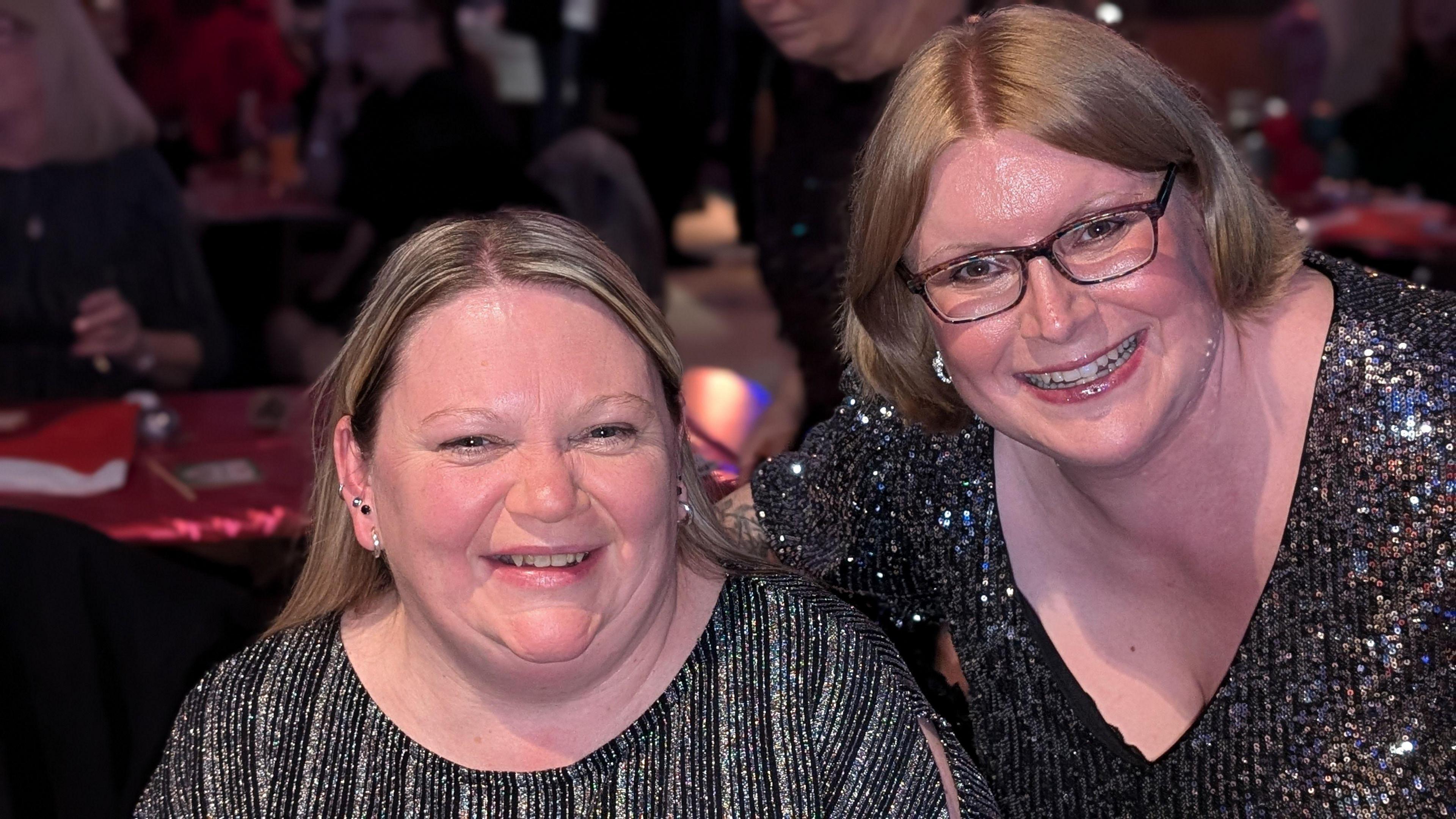 Two women wearing sparkly party tops look towards the camera and smile. Alison, to the left has blond hair and small ear-rings. Laura, to the right, has ginger-blond hair and wears glasses. Tables, decorated for Christmas, can be seen in the background