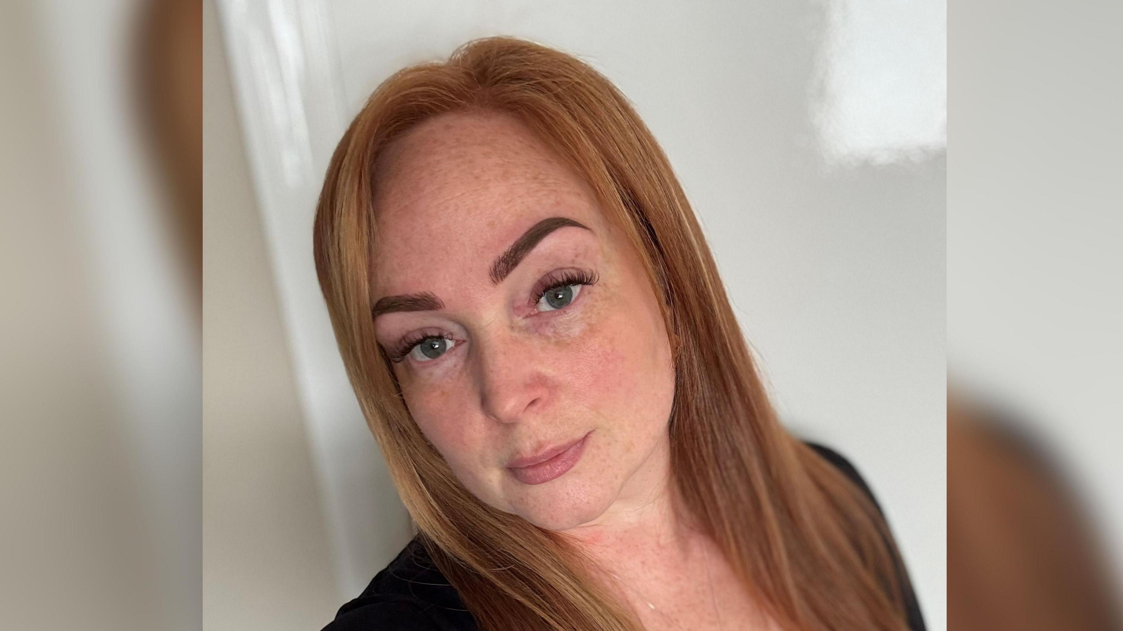 Rebecca Tyson looks at the camera in a selfie. She has long straight, reddish hair and is wearing a black top.