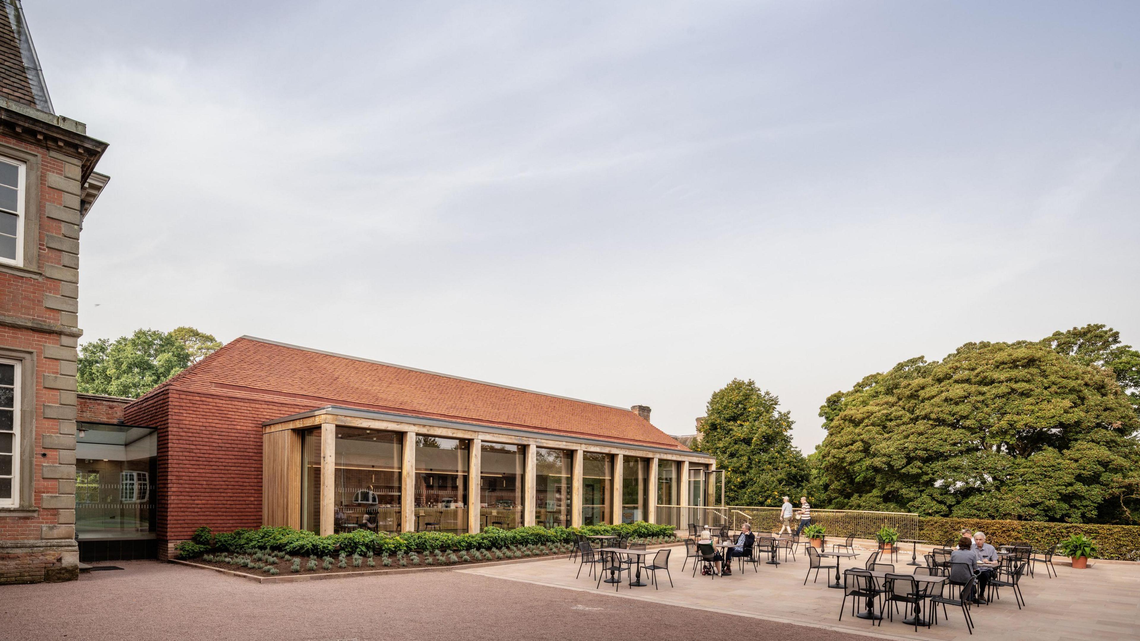 A courtyard with black tables and chairs as well as wooden and red brick building on the left. There are trees in the background.