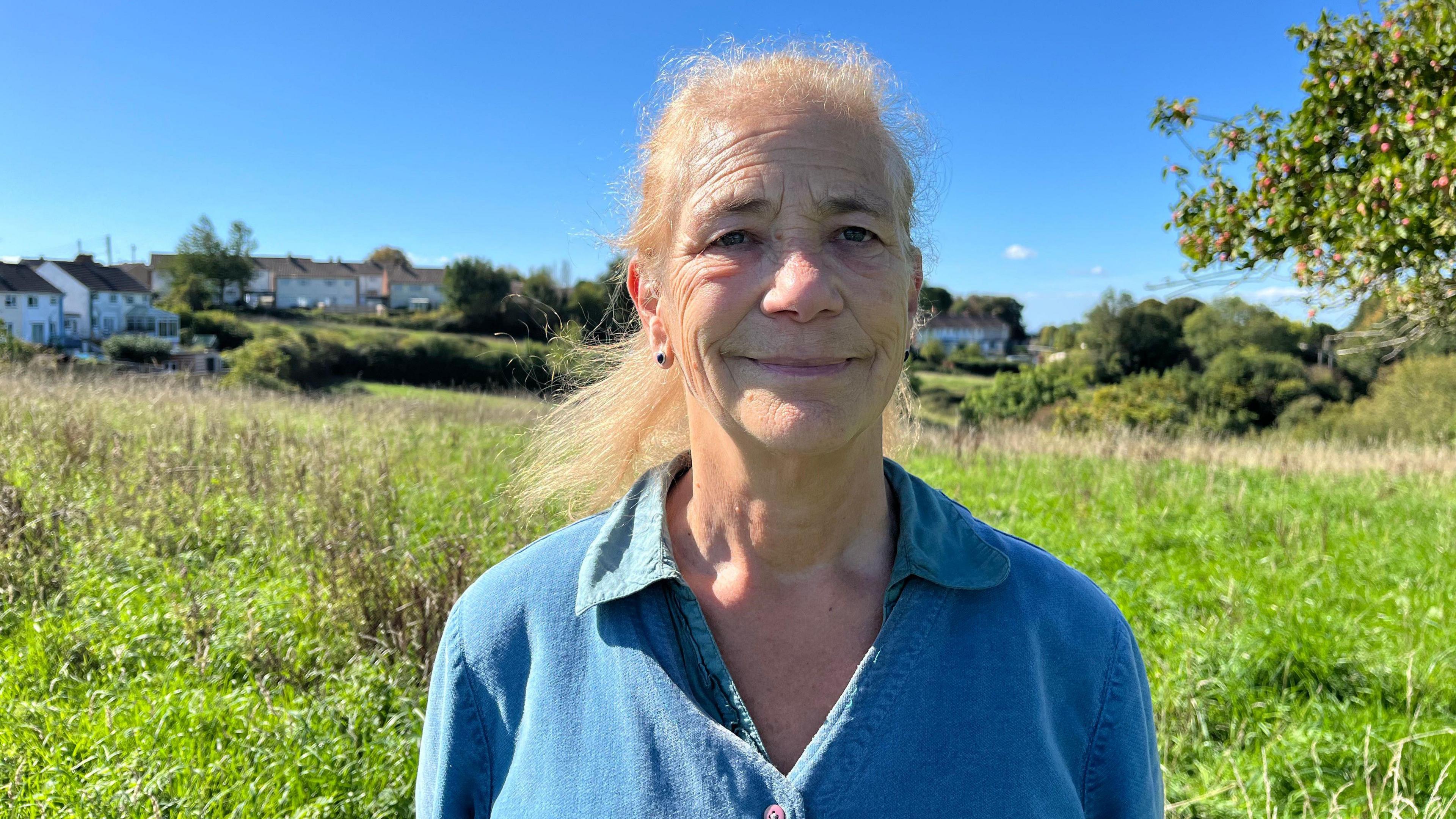 A woman in a blue top pictured from the chest up. She's looking at the camera and has a smile on her face. Behind her is a grassy orchard, and beyond that, houses.