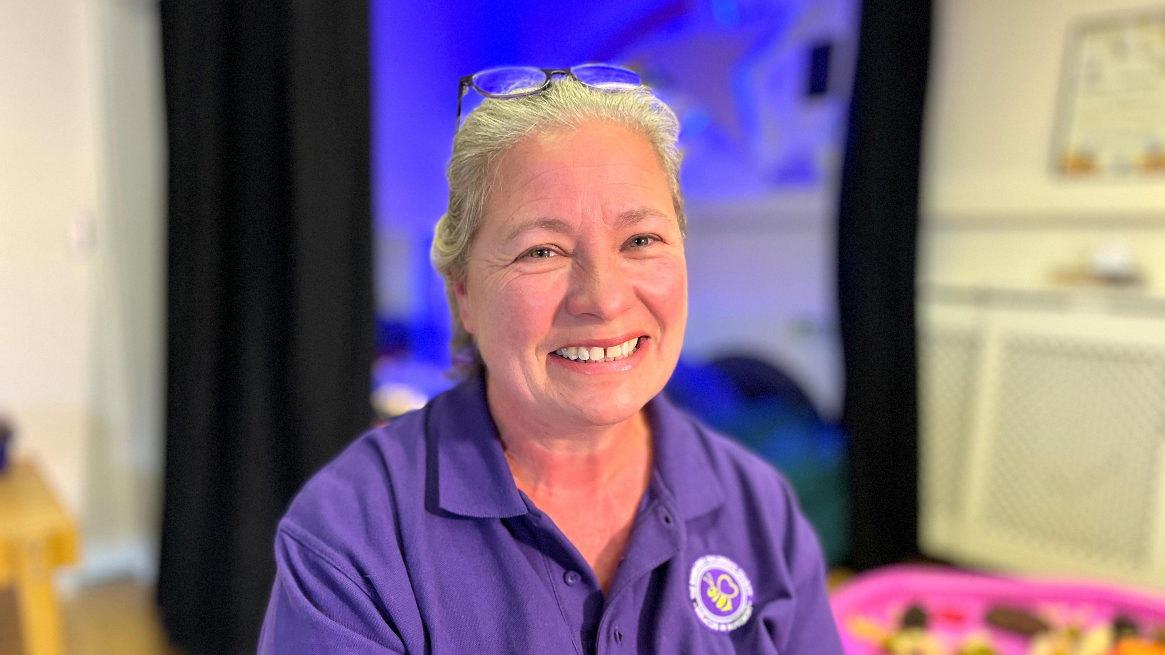 A smiling women with short blonde hair. She has glasses on her head and is wearing a purple polo shirt.